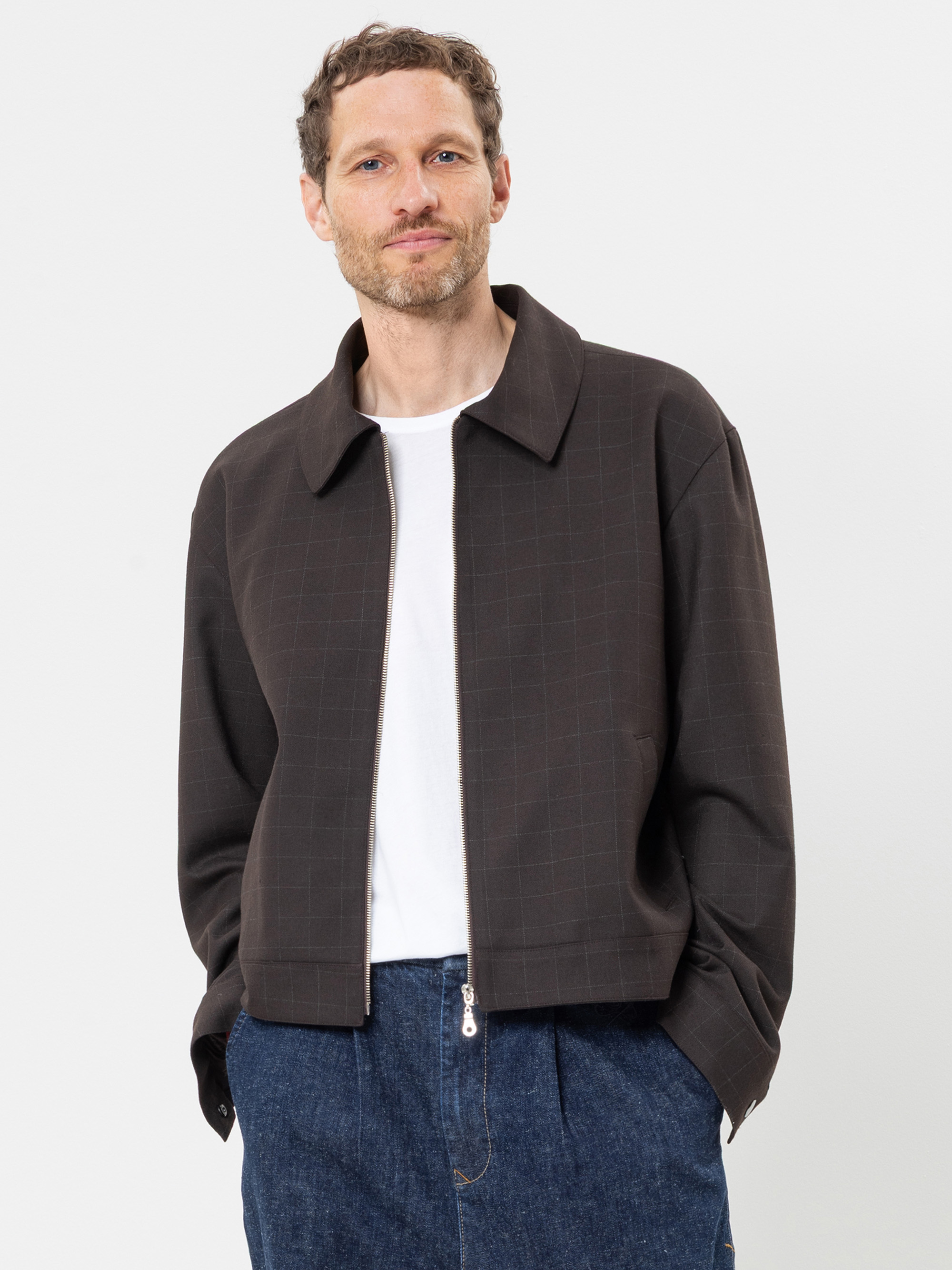 Ein Mann mit kurzen Locken und Bart trägt eine dunkle Jacke mit dezentem Karomuster, ein weißes T-Shirt und blaue Jeans. Er steht vor einem schlichten weißen Hintergrund, blickt in die Kamera und hat die Hände in den Taschen.