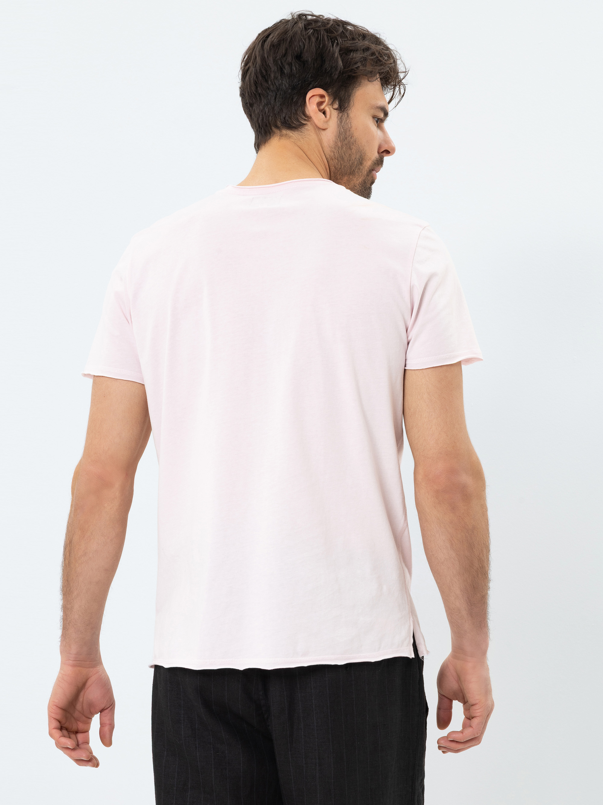 Ein Mann mit kurzen dunklen Haaren steht mit dem Gesicht zur Kamera, trägt ein hellrosa kurzärmeliges T-Shirt und eine schwarze Hose vor einem schlichten hellen Hintergrund.