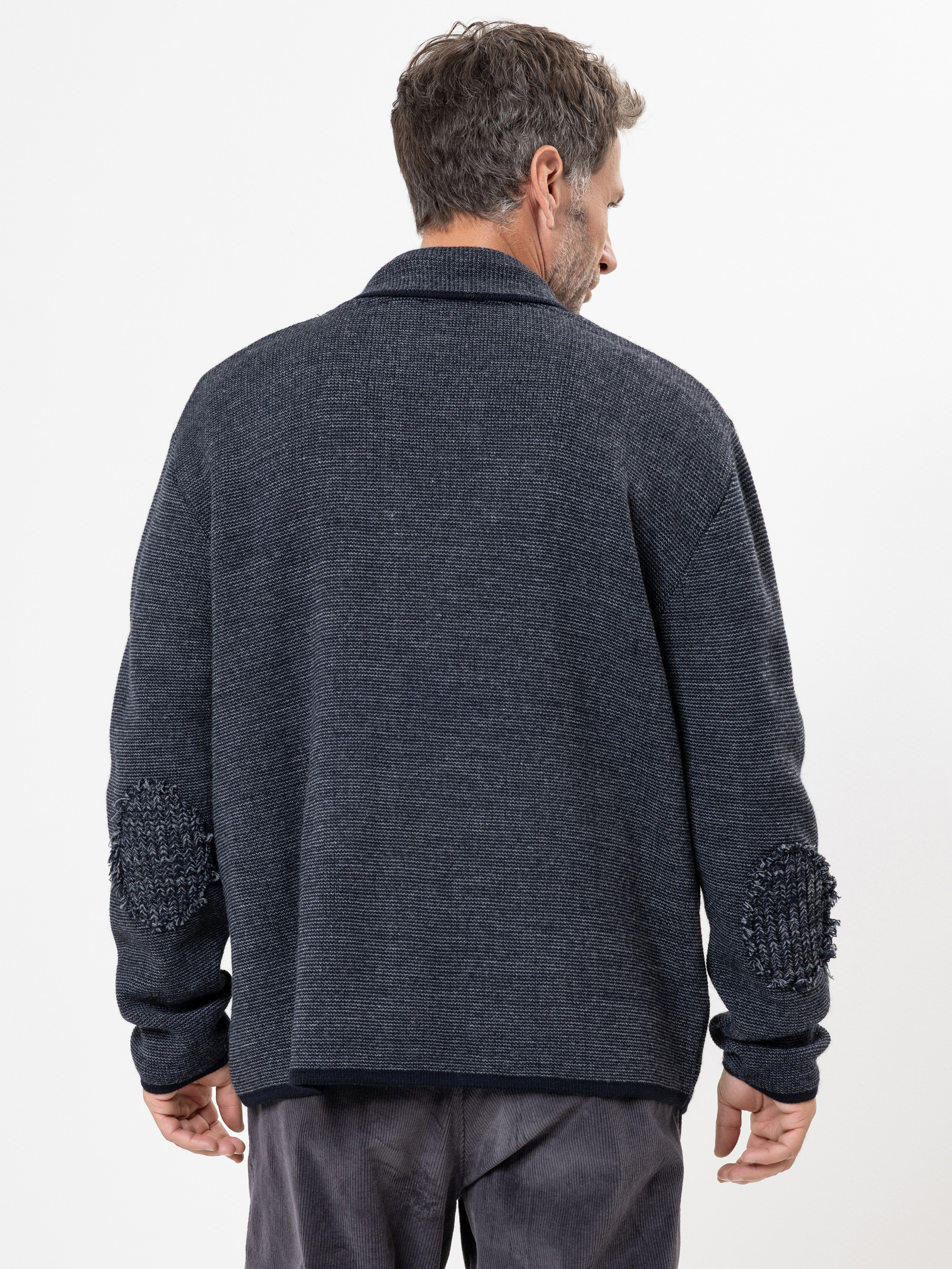 Ein Mann steht mit dem Gesicht zur Wand und trägt eine dunkelblaue, strukturierte Strickjacke mit Aufnähern an den Ellbogen und eine graue Hose vor einem schlichten weißen Hintergrund.