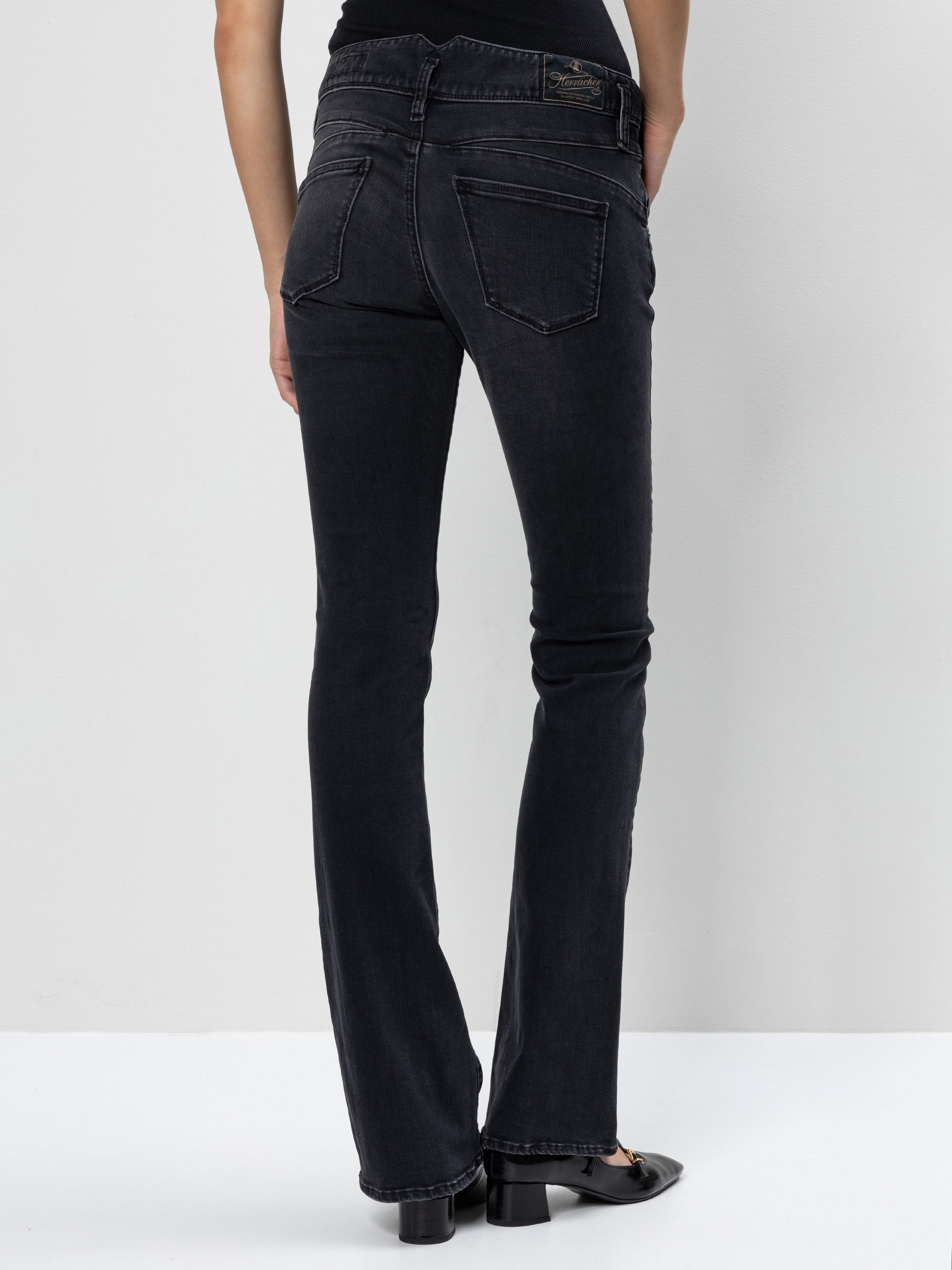 Eine Person steht von der Kamera abgewandt und trägt eine schwarze Bootcut-Jeans und schwarze Schuhe. Die Jeans hat zwei Gesäßtaschen und einen Bund mit Gürtelschlaufen. Der Hintergrund ist einfarbig und hell gefärbt.