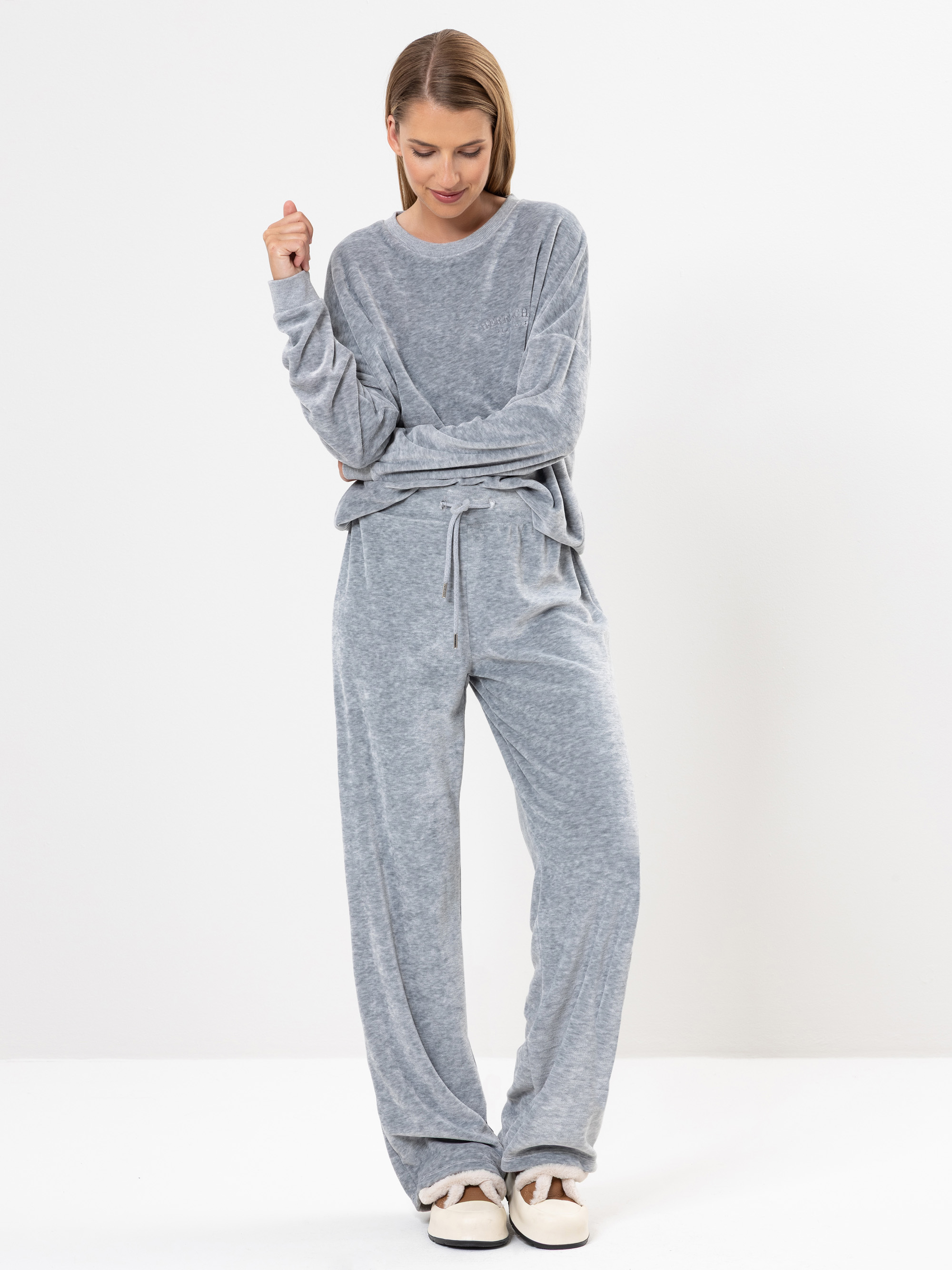 Eine Frau steht vor einem schlichten weißen Hintergrund. Sie trägt ein locker sitzendes, hellgraues Lounge-Set mit langen Ärmeln und einer Hose mit Kordelzug, dazu cremefarbene Hausschuhe. Sie hat glattes, schulterlanges Haar, blickt nach unten und lä