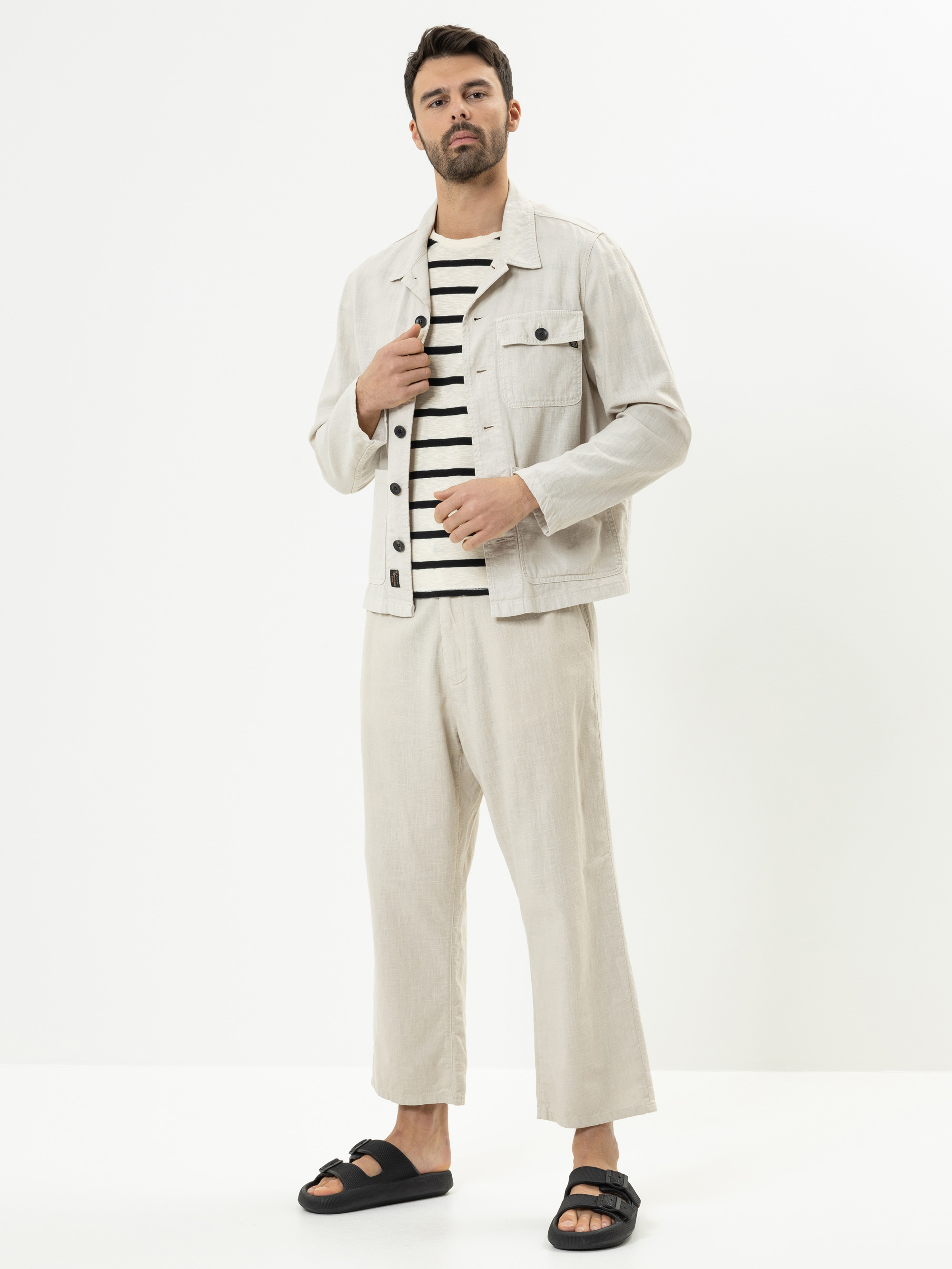 Ein Mann steht vor einem weißen Hintergrund und trägt eine hellbeige Jacke und eine passende Hose, ein schwarz-weiß gestreiftes Hemd und schwarze Sandalen. Er hat kurzes dunkles Haar und einen Bart. Mit einer Hand rückt er seine Jacke zurecht.