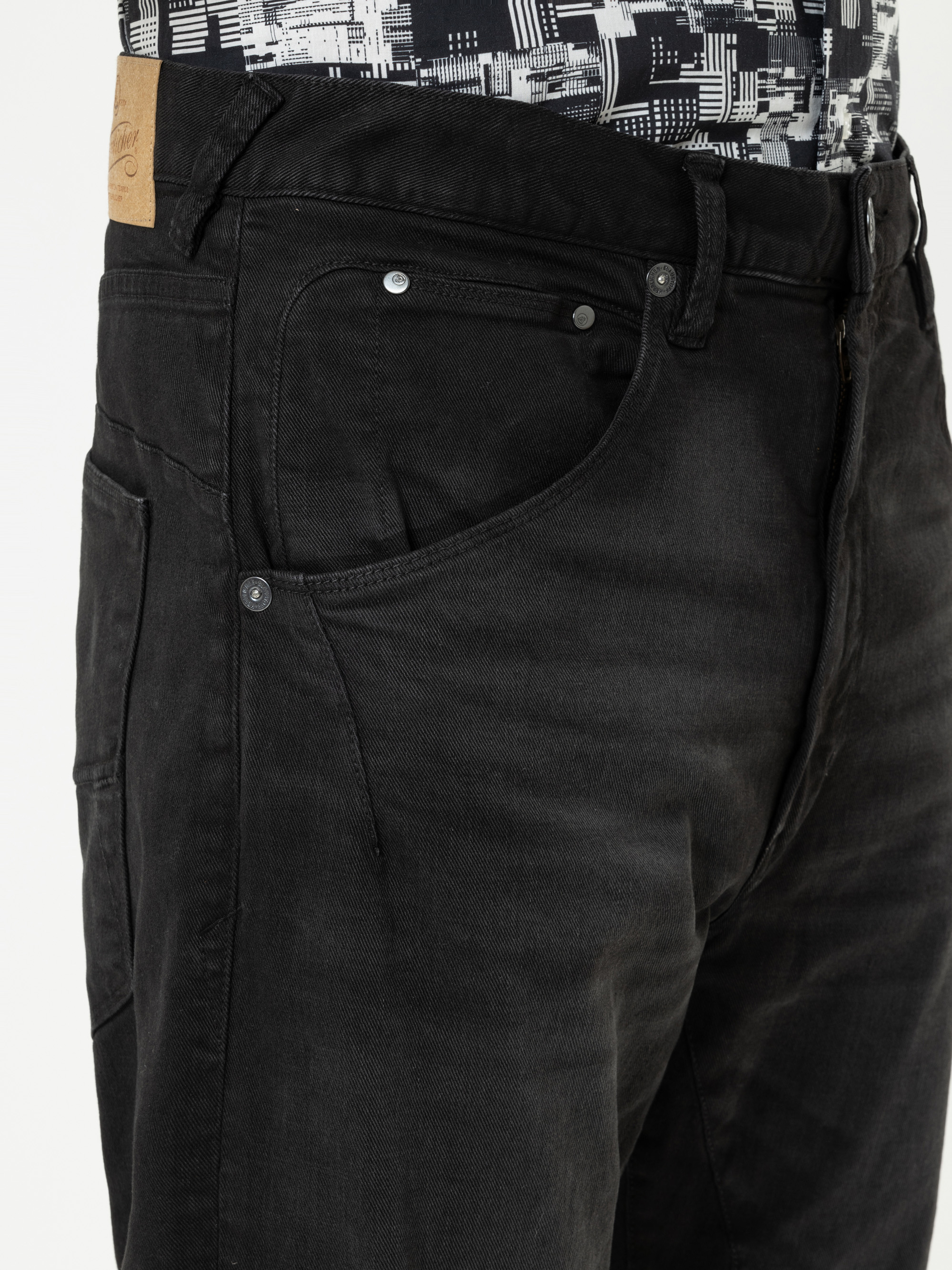 Nahaufnahme einer Person, die eine schwarze Jeans und ein gemustertes schwarz-weißes Hemd trägt. Die Jeans hat Vordertaschen mit Metallnieten, sichtbare Nähte und einen braunen Lederaufnäher am Bund.