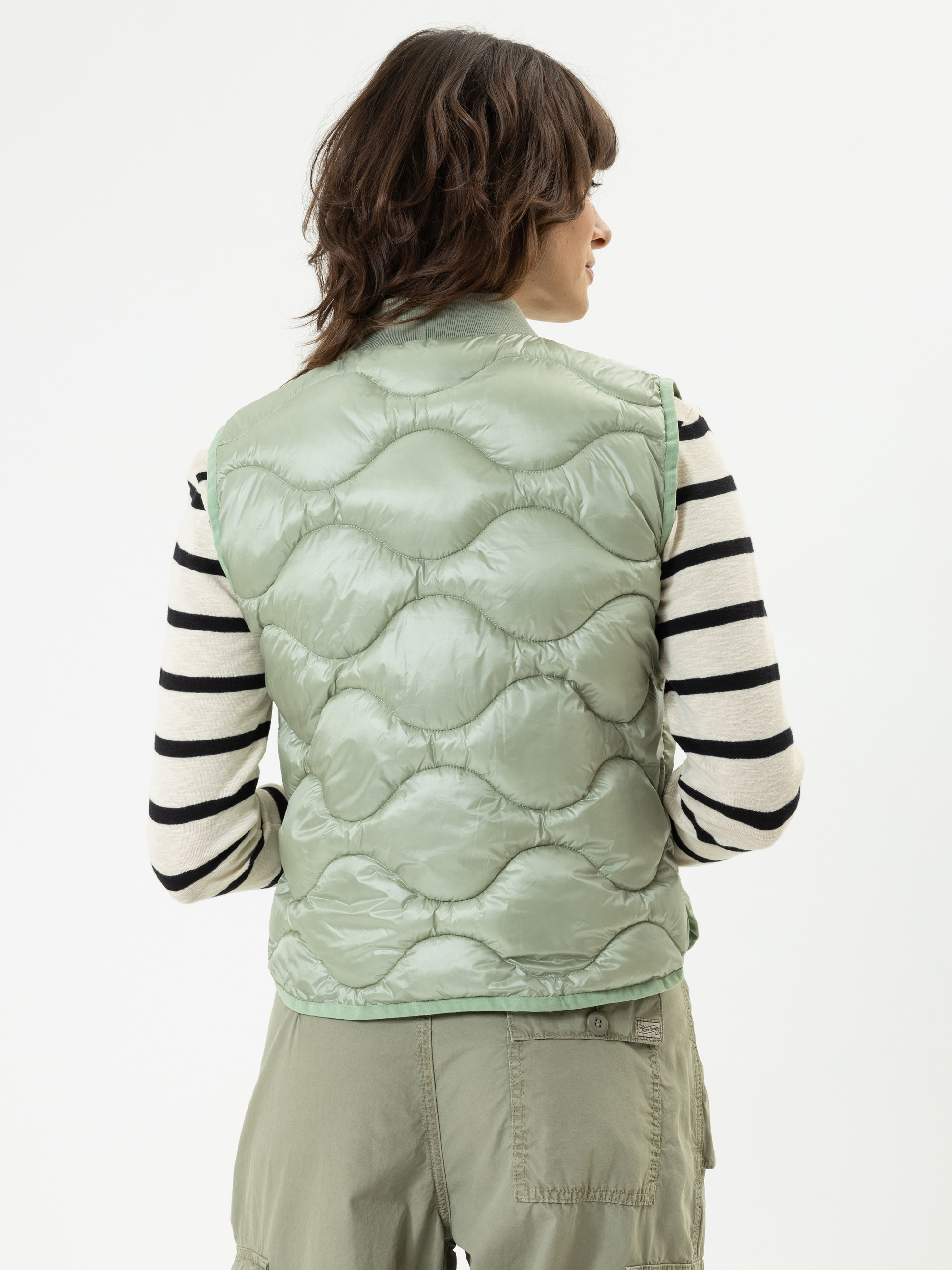 Eine Person mit schulterlangen braunen Haaren, von hinten gesehen, trägt eine hellgrüne Steppweste über einem schwarz-weiß gestreiften langärmeligen Hemd und eine grüne Cargohose vor einem schlichten weißen Hintergrund.