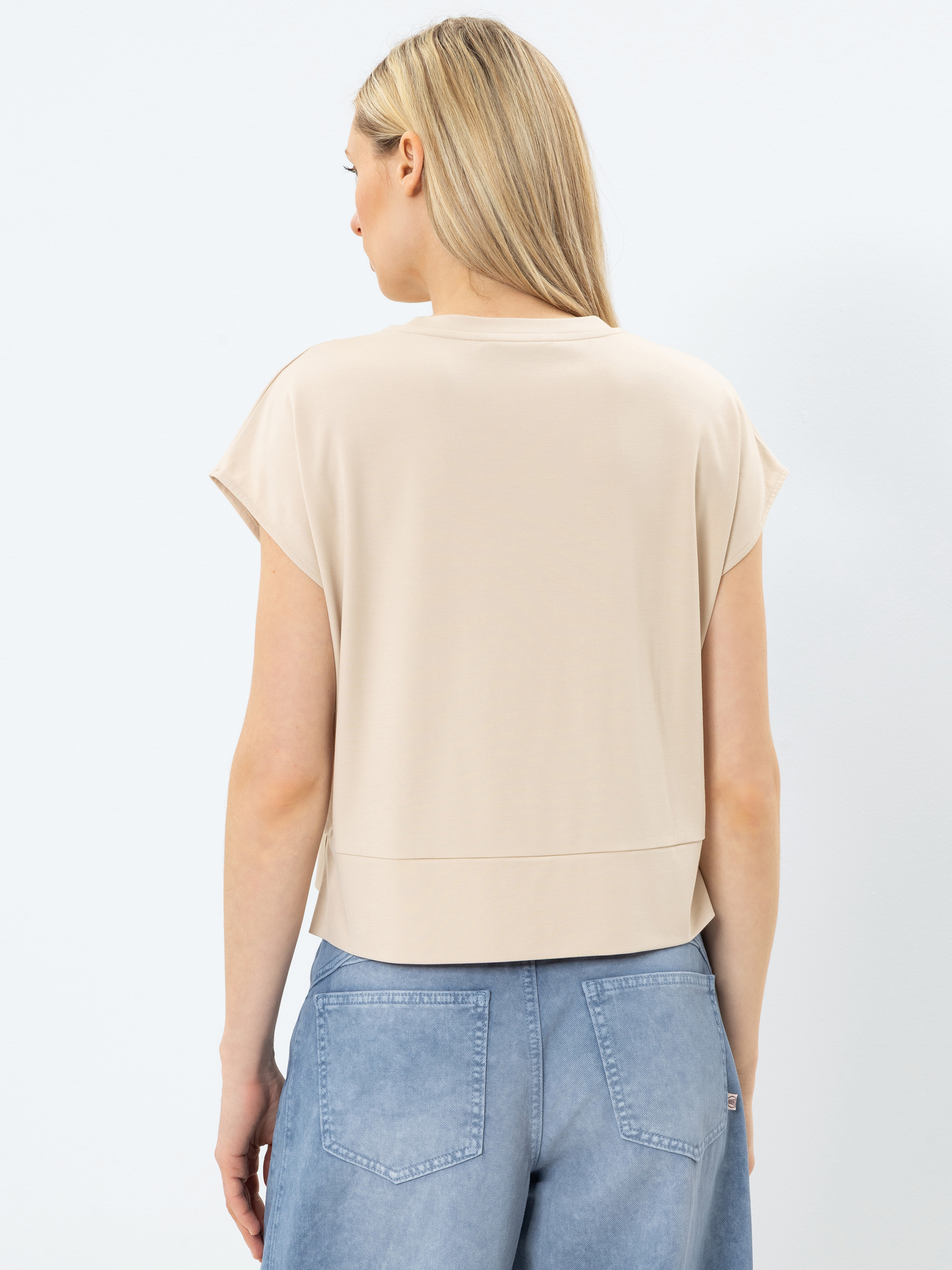 Eine Frau mit langen blonden Haaren wird von hinten gezeigt. Sie trägt ein lockeres beigefarbenes kurzärmeliges Oberteil und hellblaue Jeans vor einem schlichten weißen Hintergrund.