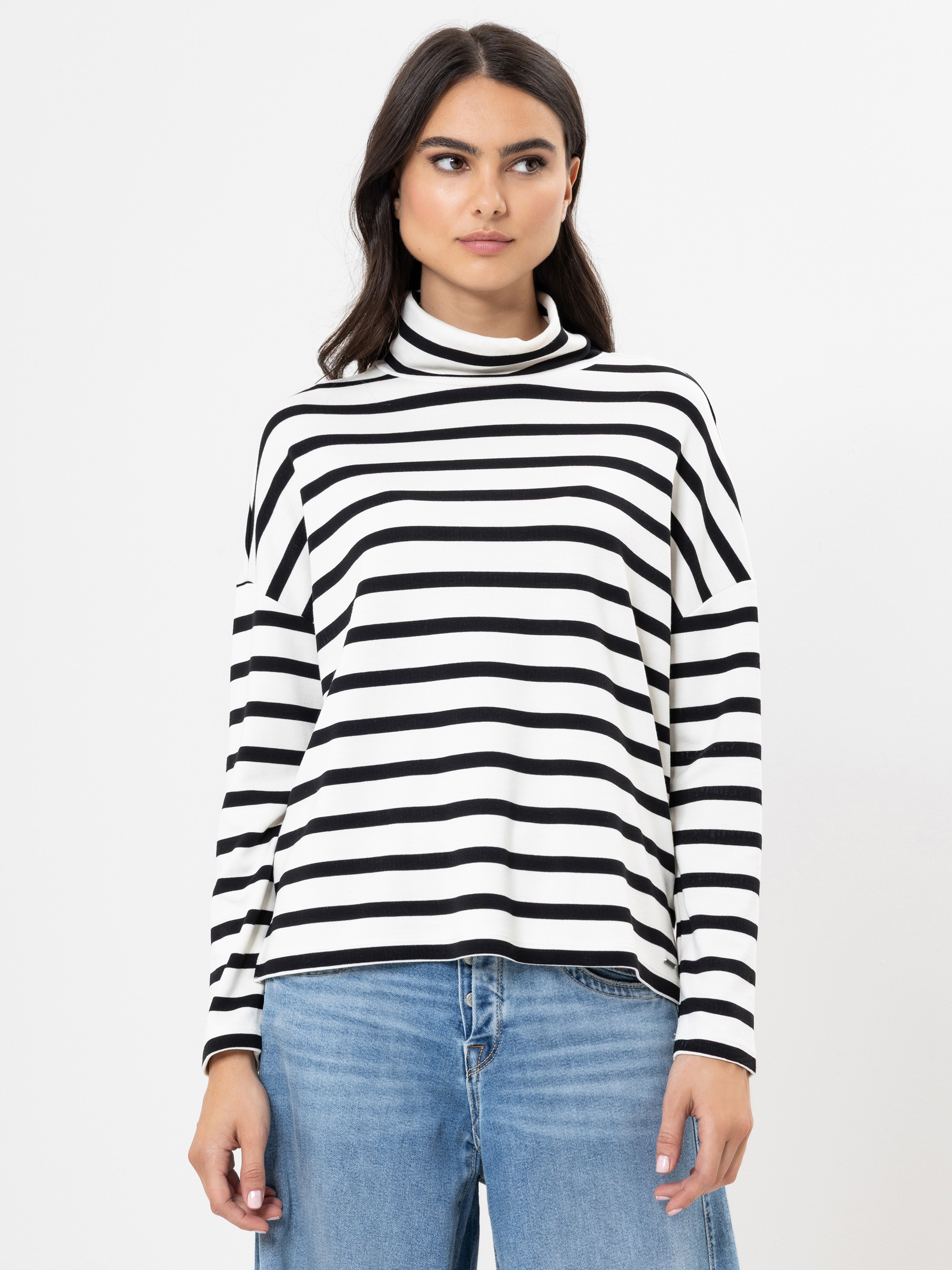 Eine Frau mit langen dunklen Haaren trägt einen schwarz-weiß gestreiften Rollkragenpullover und blaue Jeans und steht vor einem schlichten weißen Hintergrund.