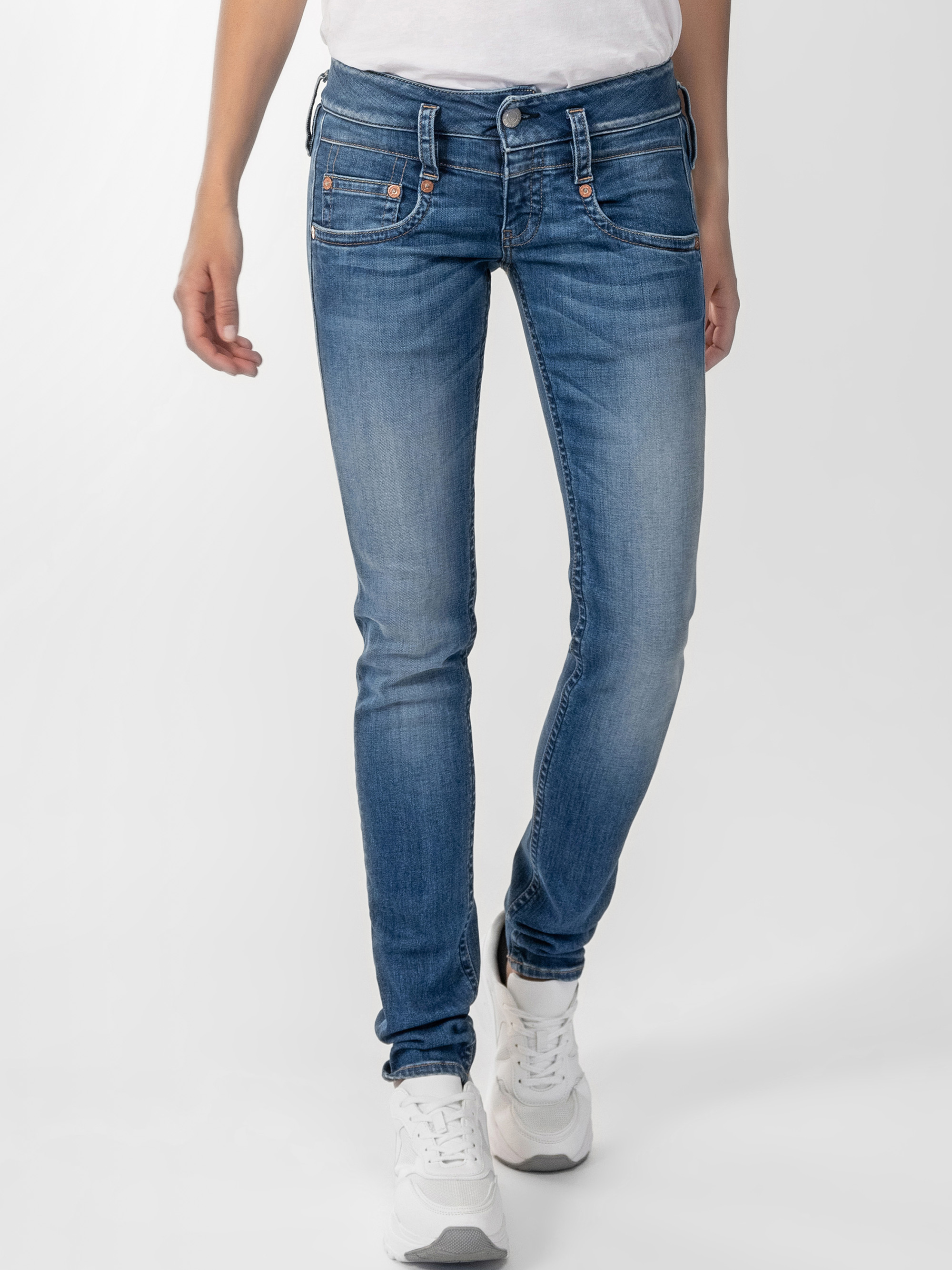Eine Person, die eine blaue Röhrenjeans, ein weißes Oberteil und weiße Turnschuhe trägt, steht vor einem schlichten hellen Hintergrund. Nur die untere Hälfte des Körpers ist sichtbar.