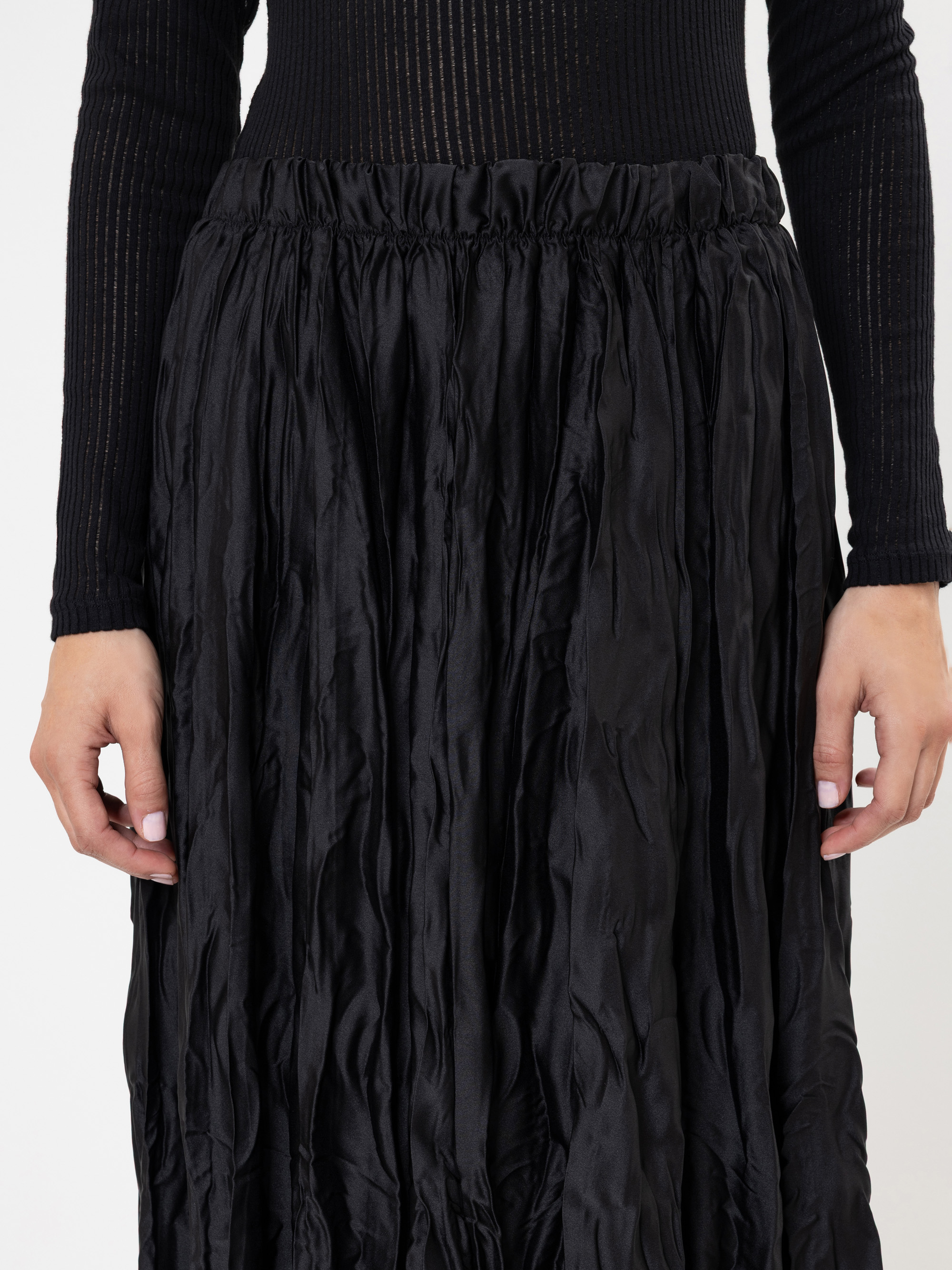 Eine Person, die ein langärmeliges, schwarzes, geripptes Oberteil und einen hochtaillierten, zerknitterten schwarzen Rock trägt, der von den Schultern bis zu den Knien reicht, wobei die Hände an der Seite ruhen.