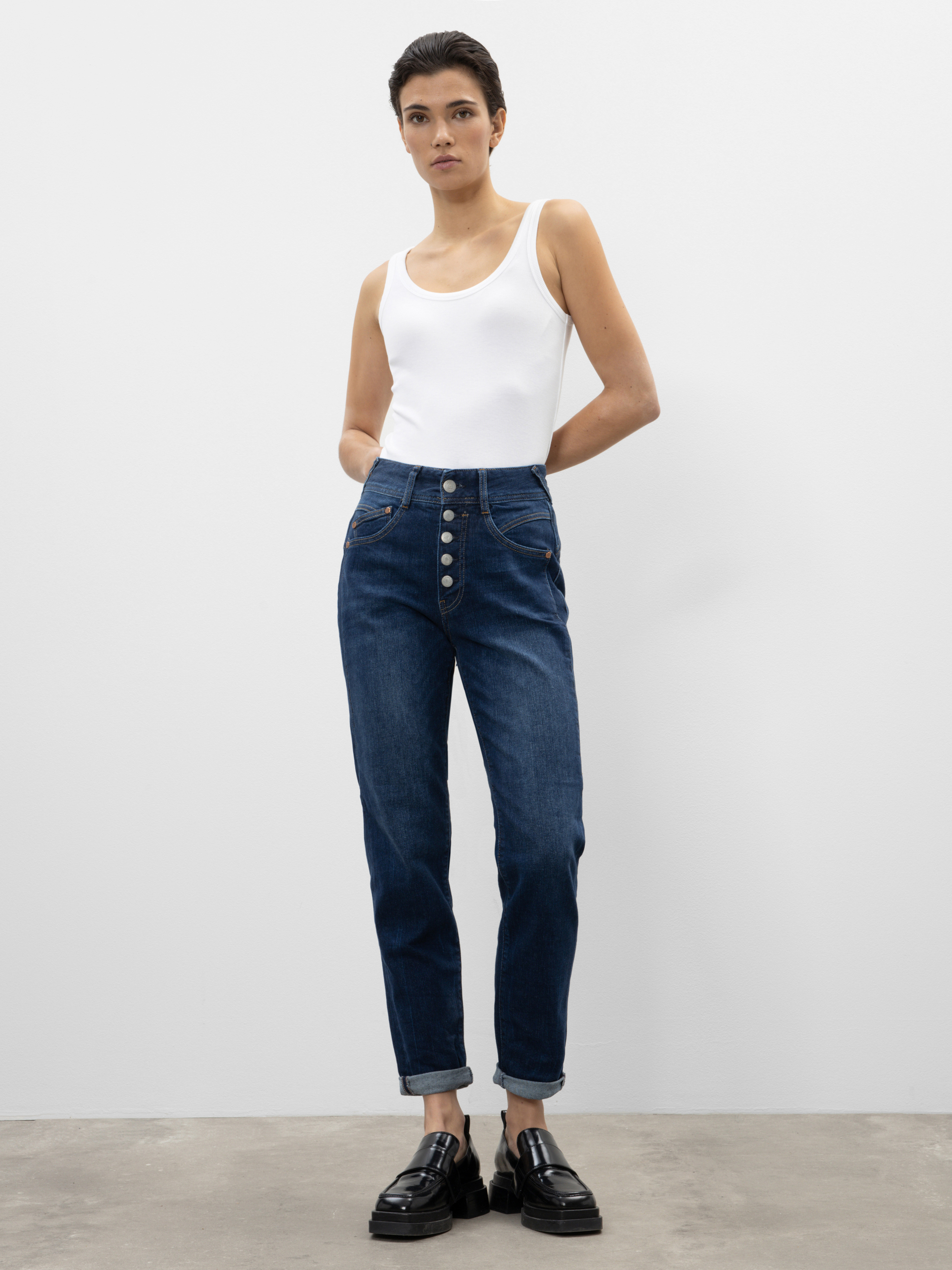 Eine Person steht vor einer schlichten weißen Wand und trägt ein weißes ärmelloses Oberteil, eine hochgekrempelte Bluejeans mit auffälligen Knöpfen und schwarze Slipper. Sie hat kurzes dunkles Haar und ihre Hände stecken in den Taschen.