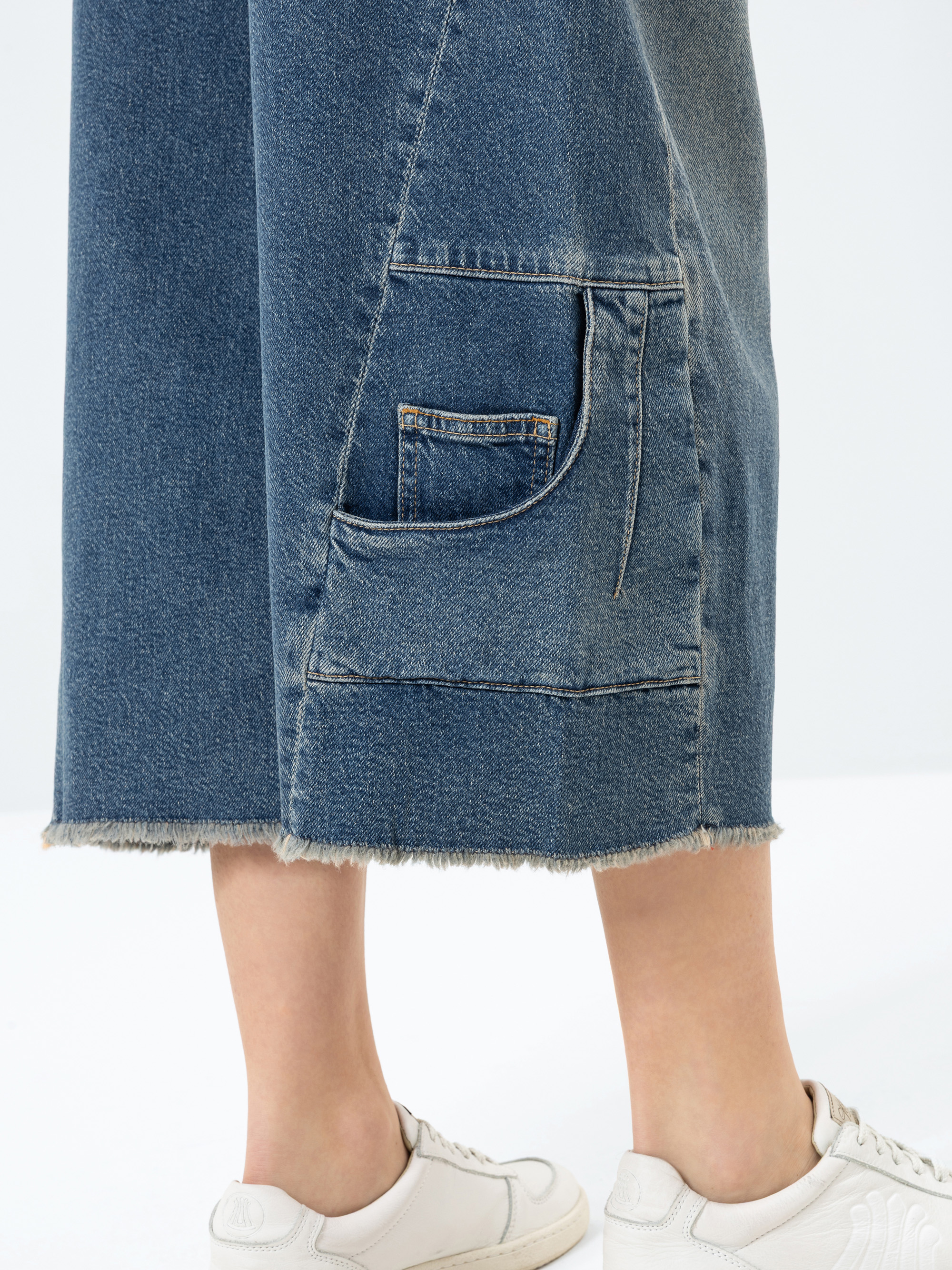 Nahaufnahme einer Person, die eine blaue Jeans-Hose mit einer großen aufgesetzten Tasche und ausgefransten Säumen trägt. Dazu trägt sie weiße Turnschuhe. Der Hintergrund ist einfarbig und hell.