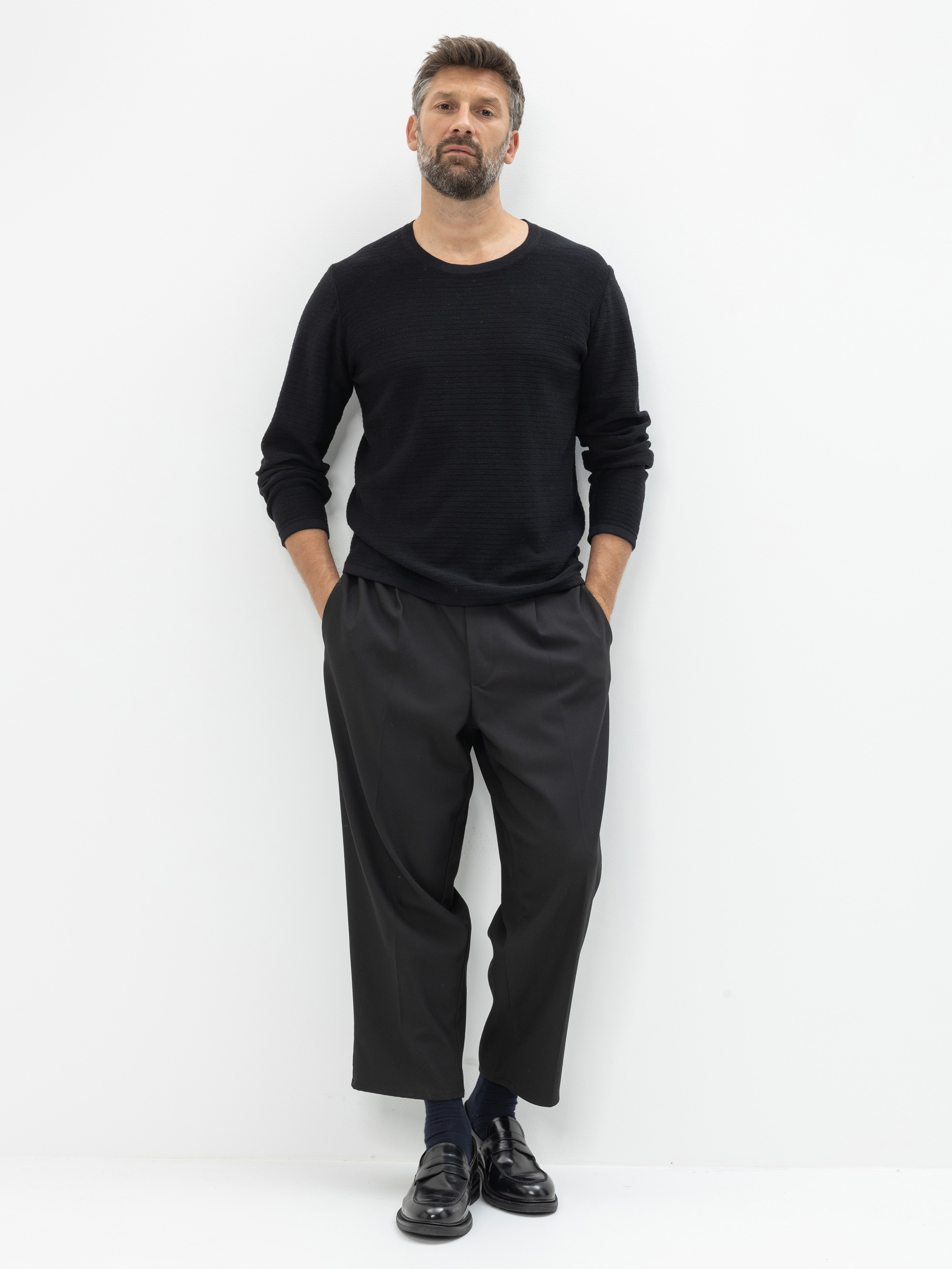 Ein Mann mit Bart steht vor einer weißen Wand, trägt ein schwarzes, langärmeliges Hemd, eine dunkle, weite Hose, marineblaue Socken und schwarze Anzugschuhe, die Hände in den Taschen.