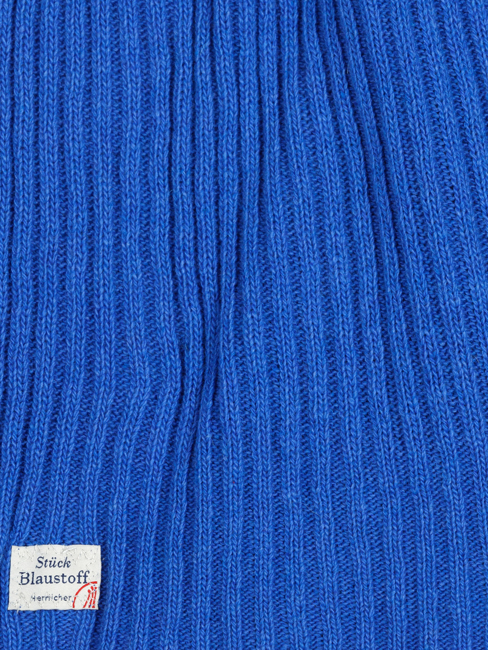 Nahaufnahme eines blauen Rippenstrickstoffs mit einem kleinen weißen Etikett in der unteren linken Ecke, das Text und ein rotes grafisches Muster enthält.