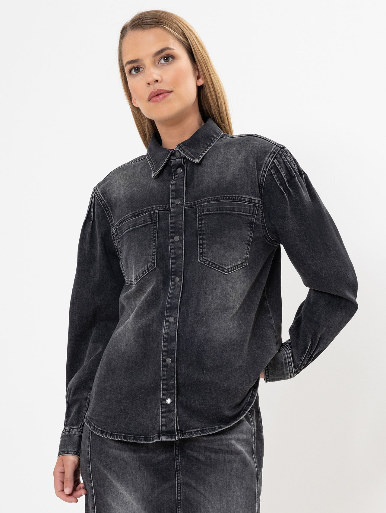 Eine Frau mit glattem blondem Haar trägt ein schwarzes, verblichenes Jeanshemd mit langen Ärmeln und zwei Brusttaschen. Sie steht vor einem schlichten weißen Hintergrund und blickt leicht zur Seite.