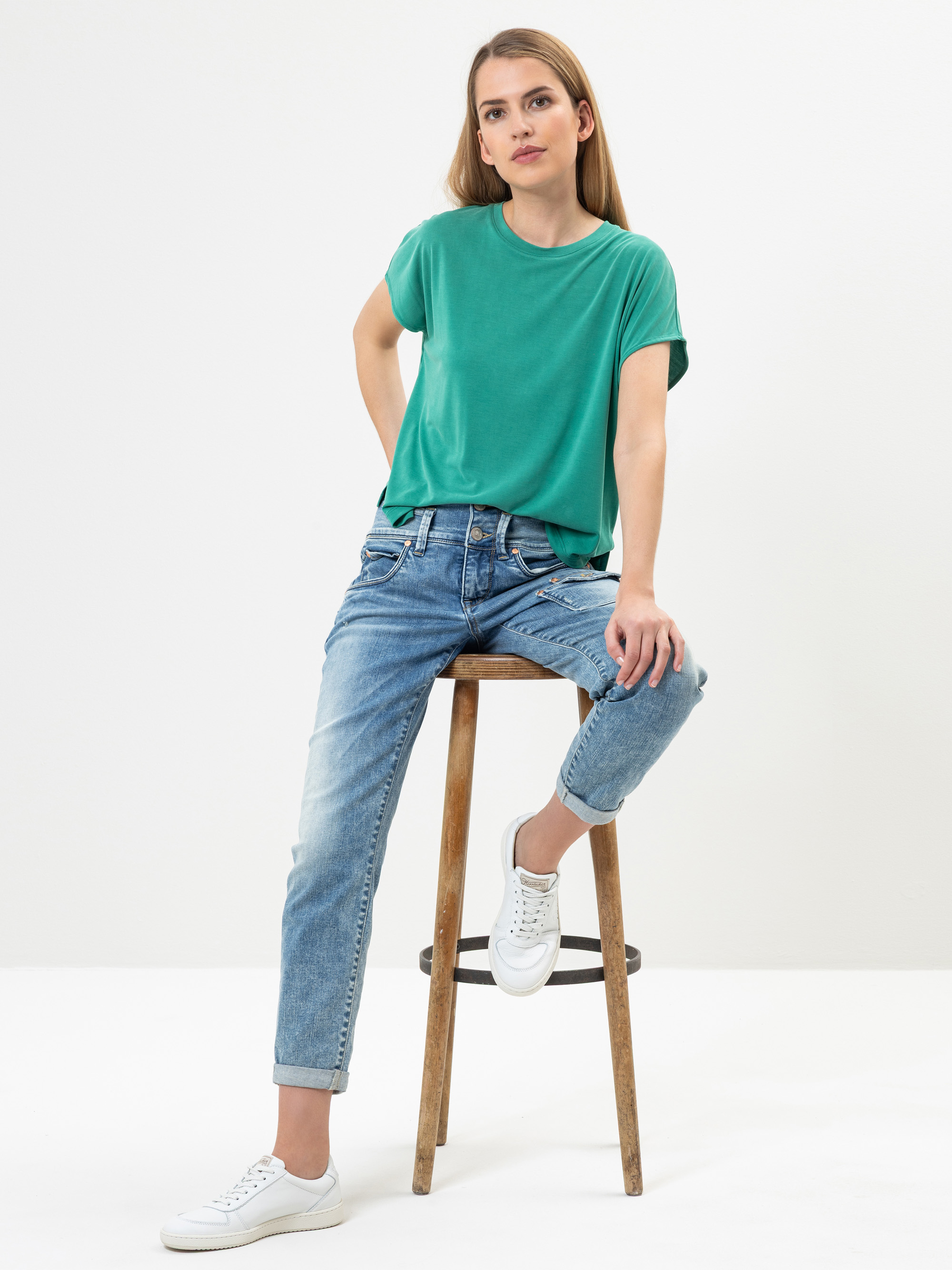 Eine Frau mit langen Haaren, die ein grünes T-Shirt, blaue Jeans und weiße Turnschuhe trägt, sitzt lässig auf einem Holzhocker vor einem schlichten weißen Hintergrund.