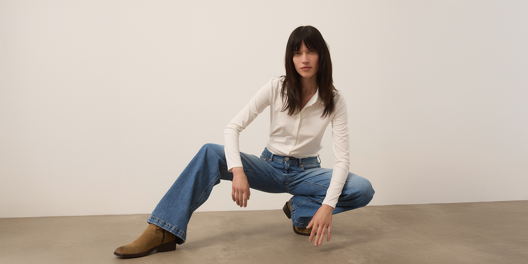 Eine Person mit langen dunklen Haaren, die ein weißes langärmeliges Hemd, blaue Jeans und braune Stiefel trägt, posiert in gebückter Haltung auf einem Betonboden vor einer schlichten weißen Wand.