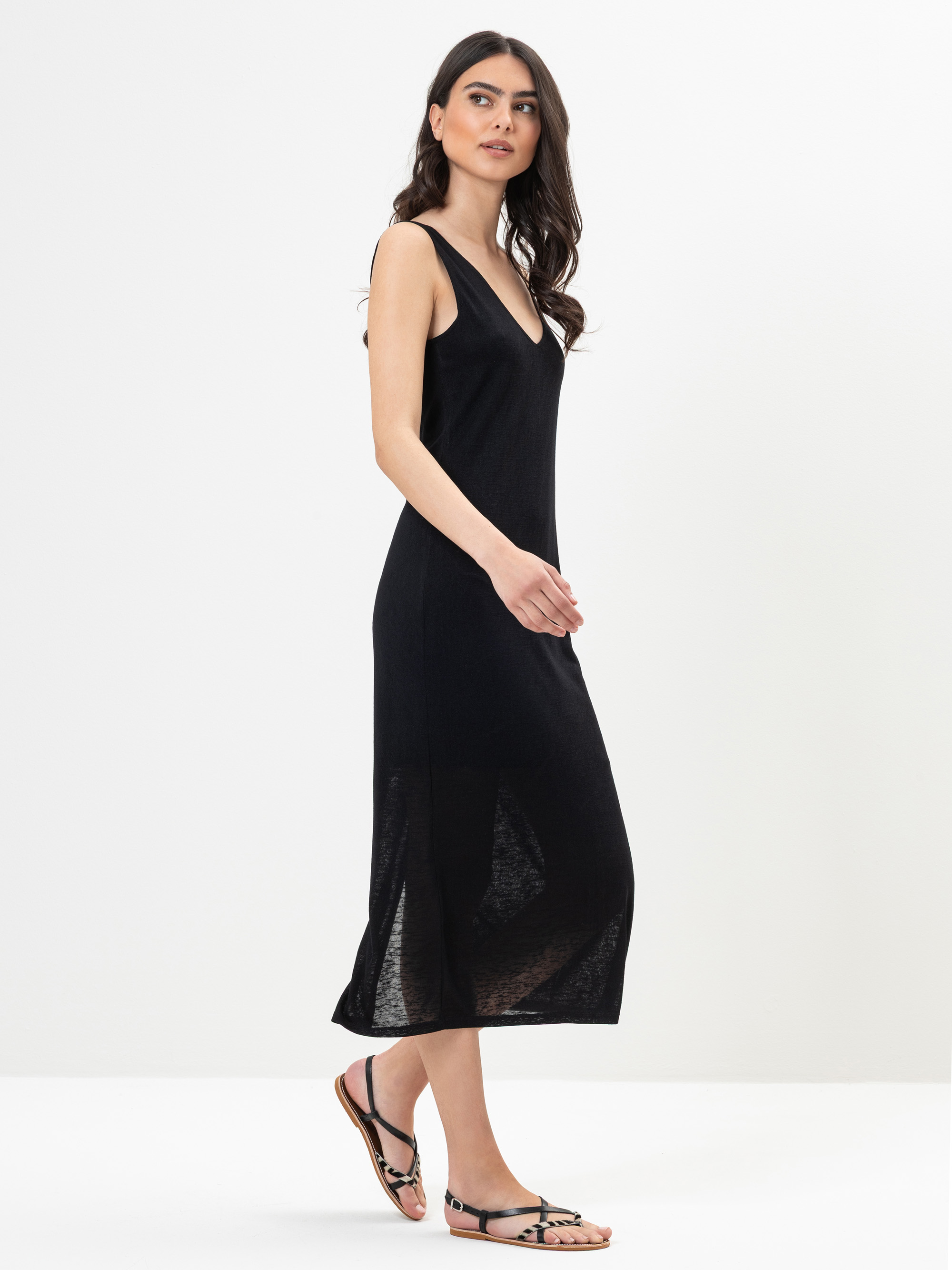 Eine Frau mit langen dunklen Haaren trägt ein ärmelloses schwarzes Midikleid und schwarze Sandalen und steht vor einem schlichten weißen Hintergrund. Sie steht seitlich und blickt leicht in die Kamera.