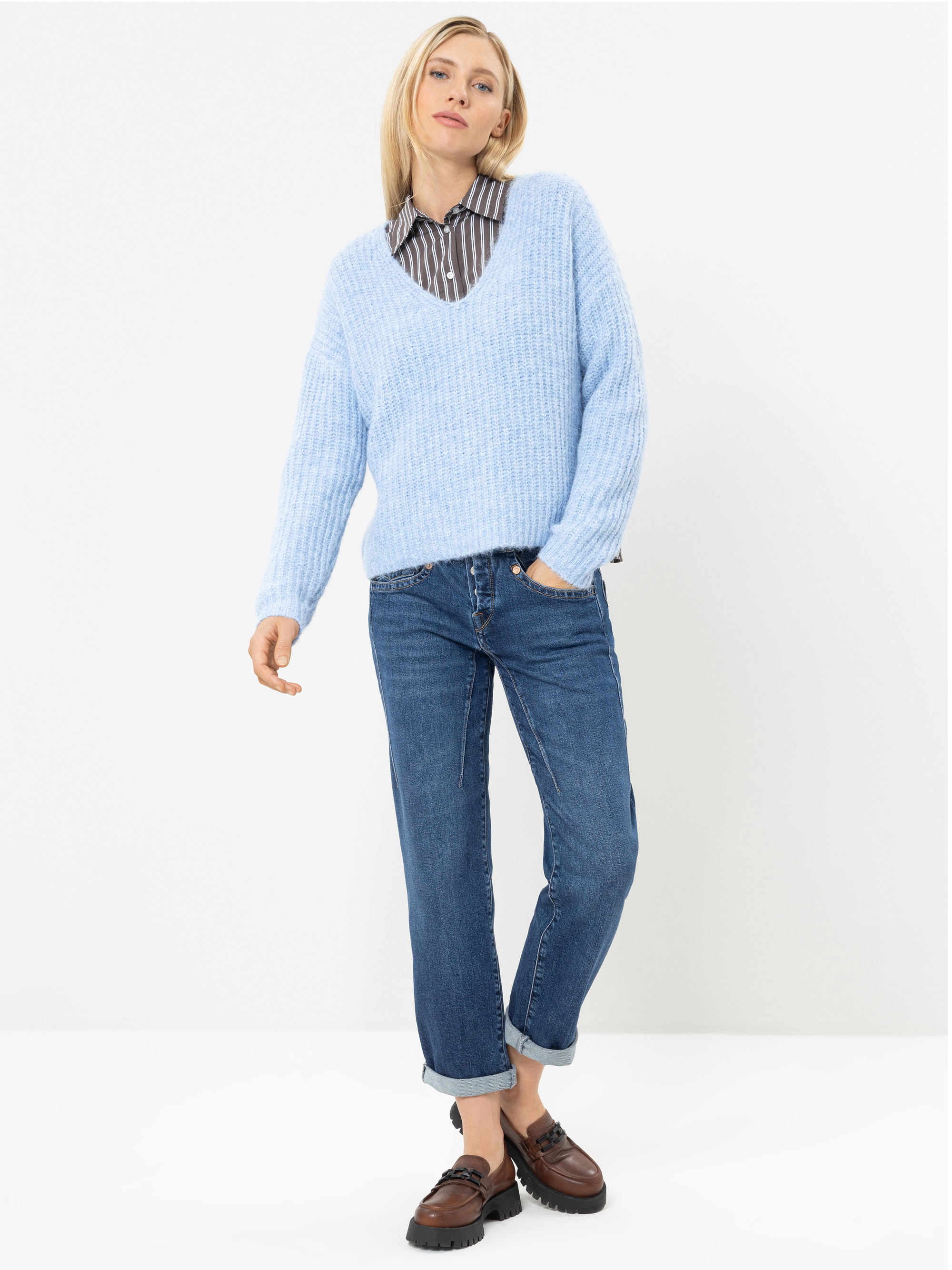 Eine Person steht vor einem weißen Hintergrund und trägt einen hellblauen Pullover mit V-Ausschnitt über einem gestreiften Hemd, eine blaue Jeans mit Umschlag und braune Schuhe. Sie hat glattes blondes Haar und eine entspannte Körperhaltung, wobei ein