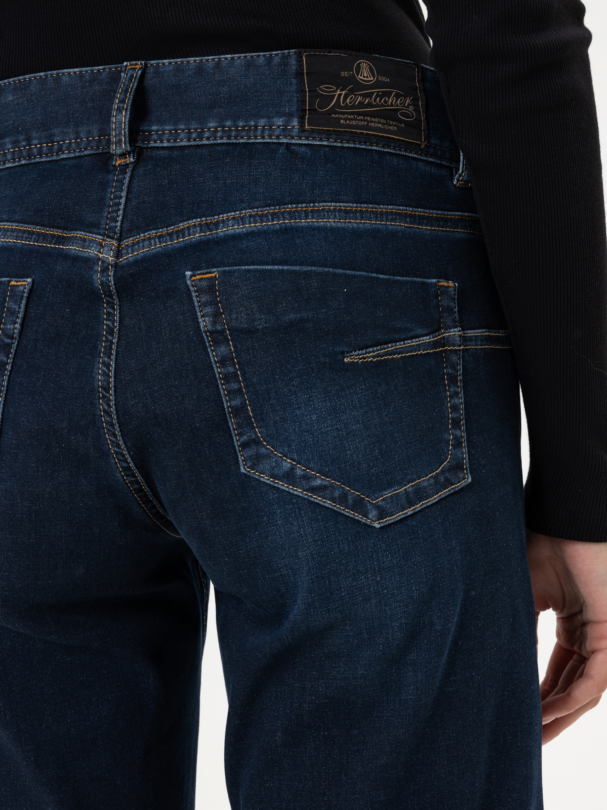 Eine Person, die eine dunkelblaue Jeans und ein schwarzes Oberteil trägt, von der Taille abwärts gezeigt. Die Jeans hat sichtbare Nähte, Gesäßtaschen und einen braunen Markenaufnäher am Bund.