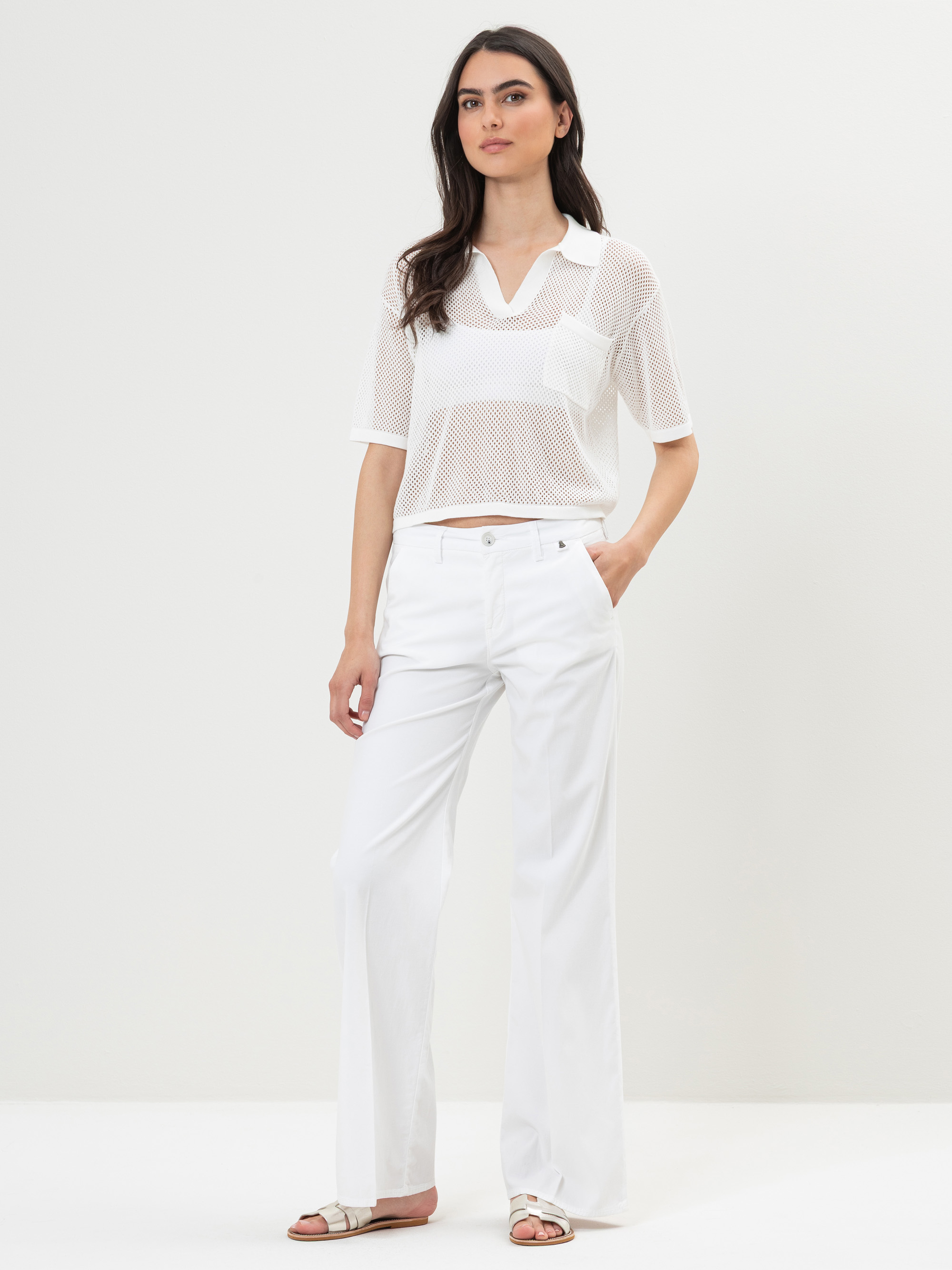 Eine Frau steht vor einem schlichten Hintergrund und trägt ein weißes, kurzärmeliges Netzoberteil mit Kragen, eine weiße Hose mit weitem Bein und weiße Sandalen. Eine Hand steckt in ihrer Tasche und sie hat einen neutralen Gesichtsausdruck.