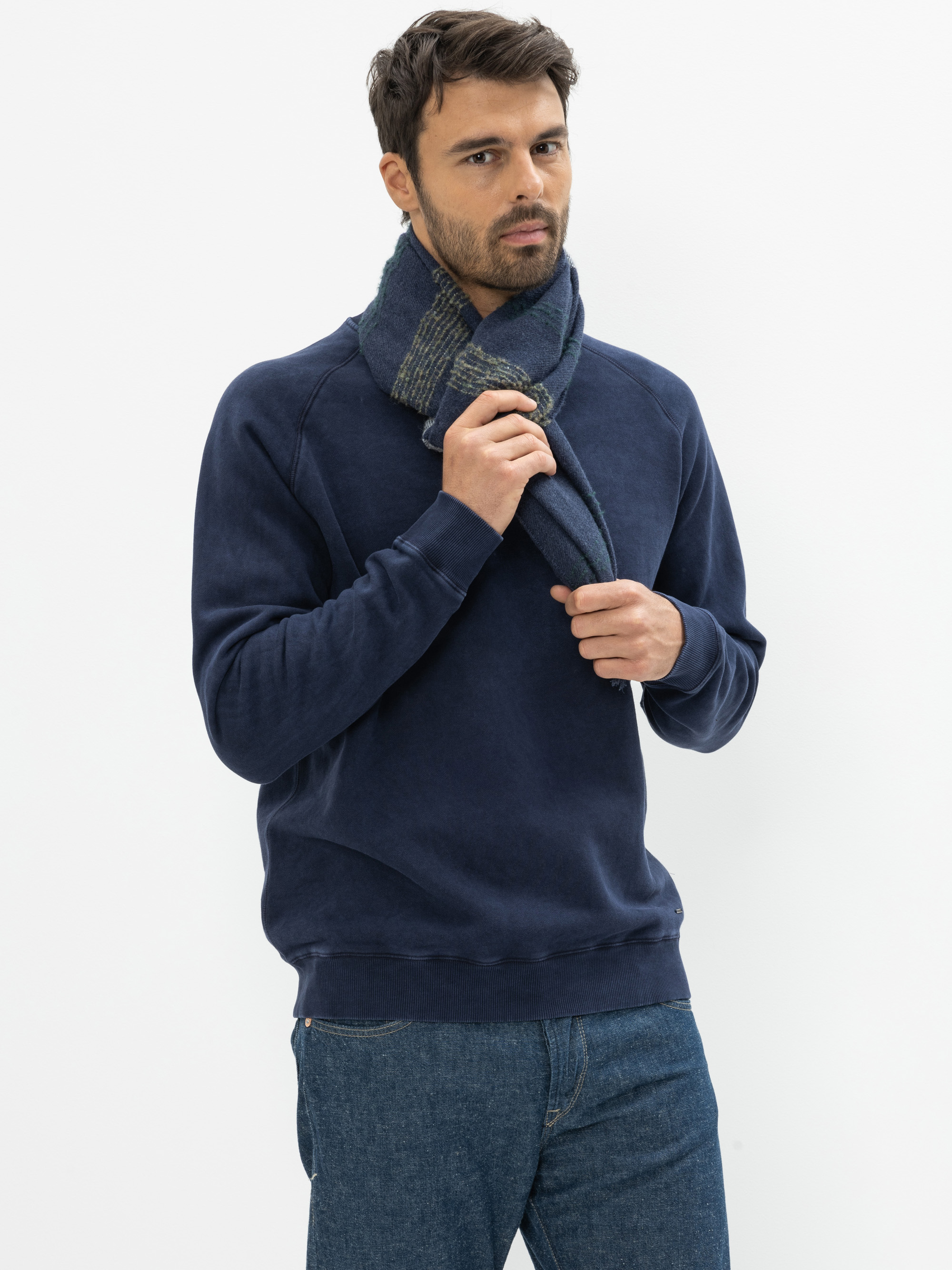 Ein Mann mit kurzen dunklen Haaren und einem Bart trägt ein marineblaues Sweatshirt, blaue Jeans und einen blau-grauen Schal um den Hals und steht vor einem schlichten weißen Hintergrund.