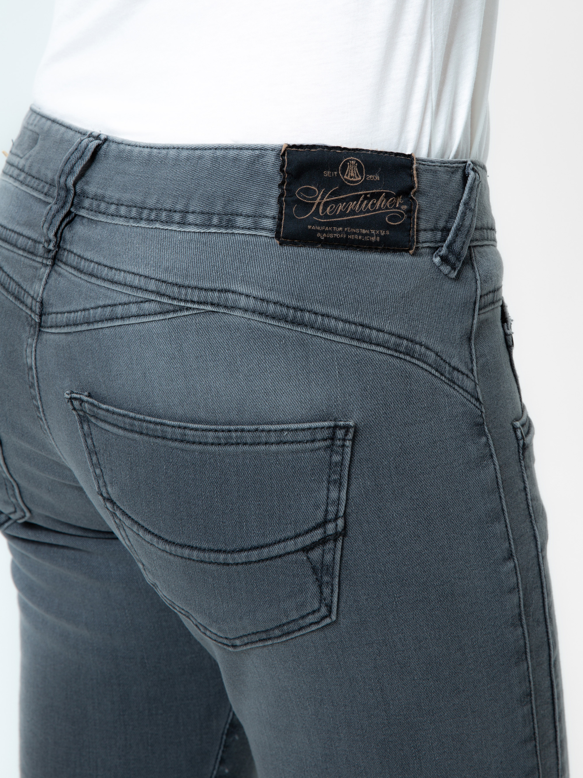 Nahaufnahme einer Person, die eine graue Jeans und ein weißes Oberteil trägt. Man sieht die Gesäßtasche und ein schwarzes Herrlicher-Markenetikett am Bund.