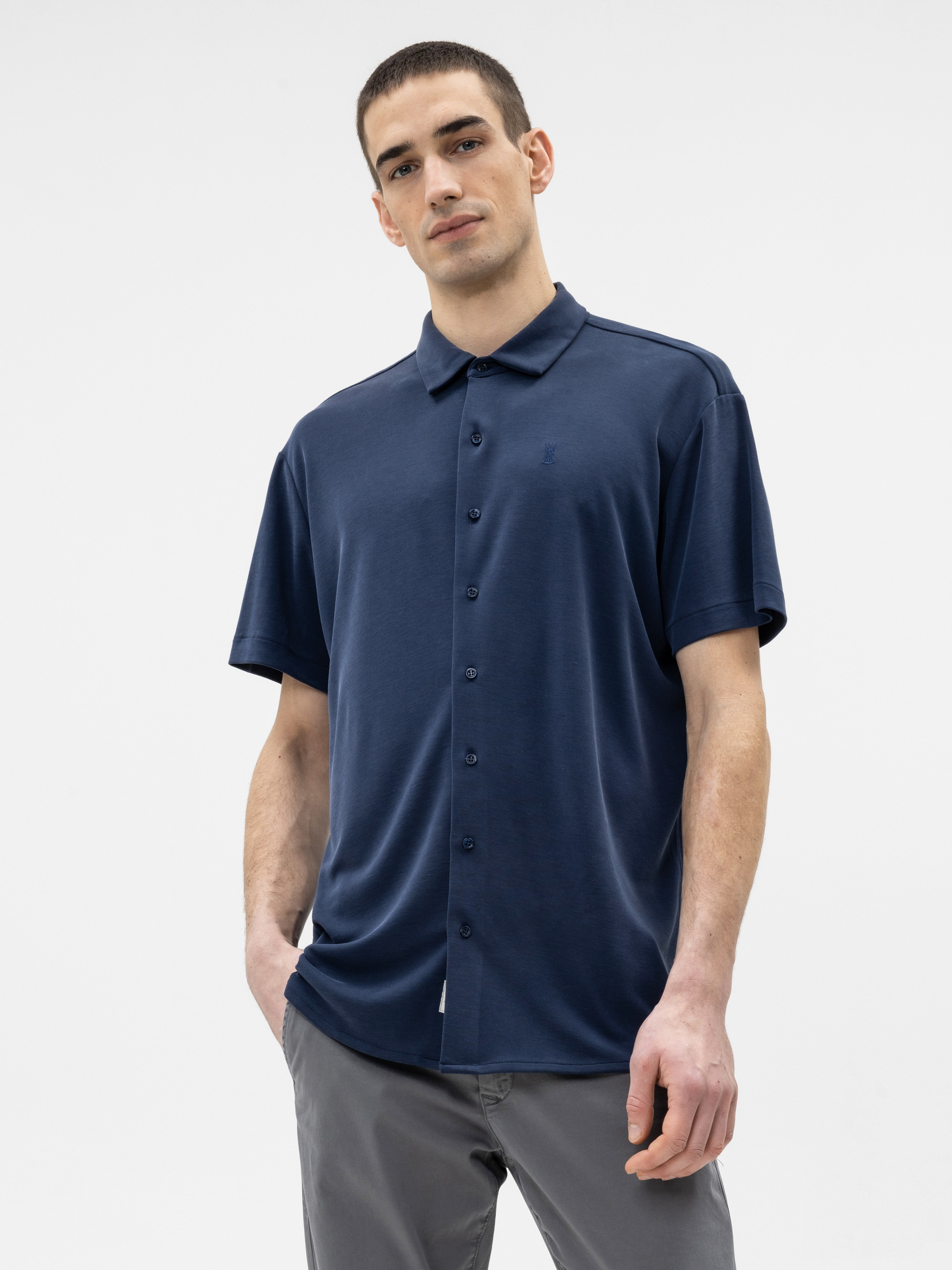 Ein Mann steht vor einem weißen Hintergrund, trägt ein dunkelblaues, kurzärmeliges Hemd und eine graue Hose, hat eine Hand in der Tasche und einen neutralen Gesichtsausdruck.