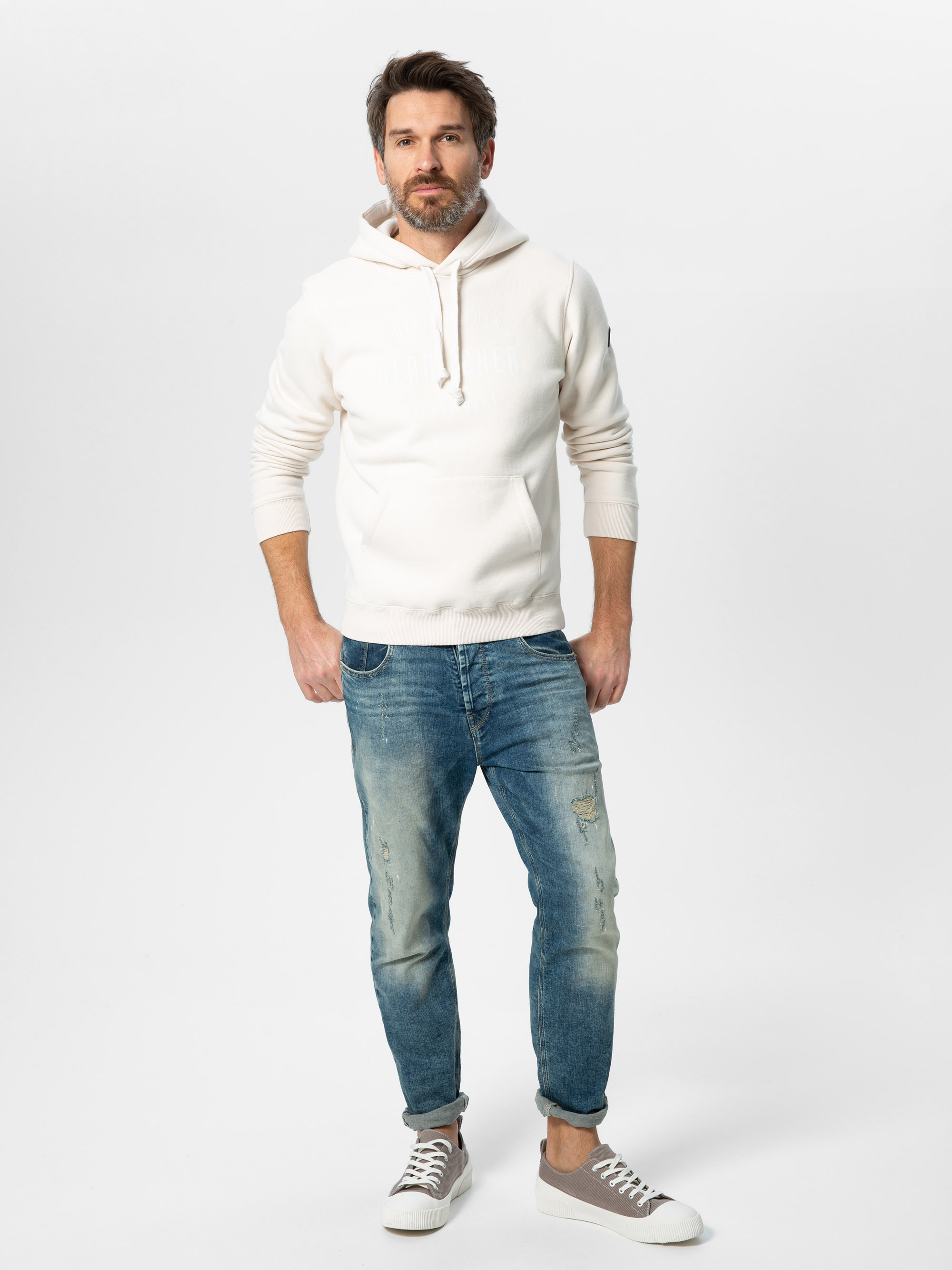 Ein bärtiger Mann steht der Kamera gegenüber und trägt einen cremefarbenen Kapuzenpullover, blaue Jeans mit Bündchen und graue Turnschuhe. Er hat eine Hand in der Tasche und steht vor einem schlichten weißen Hintergrund.