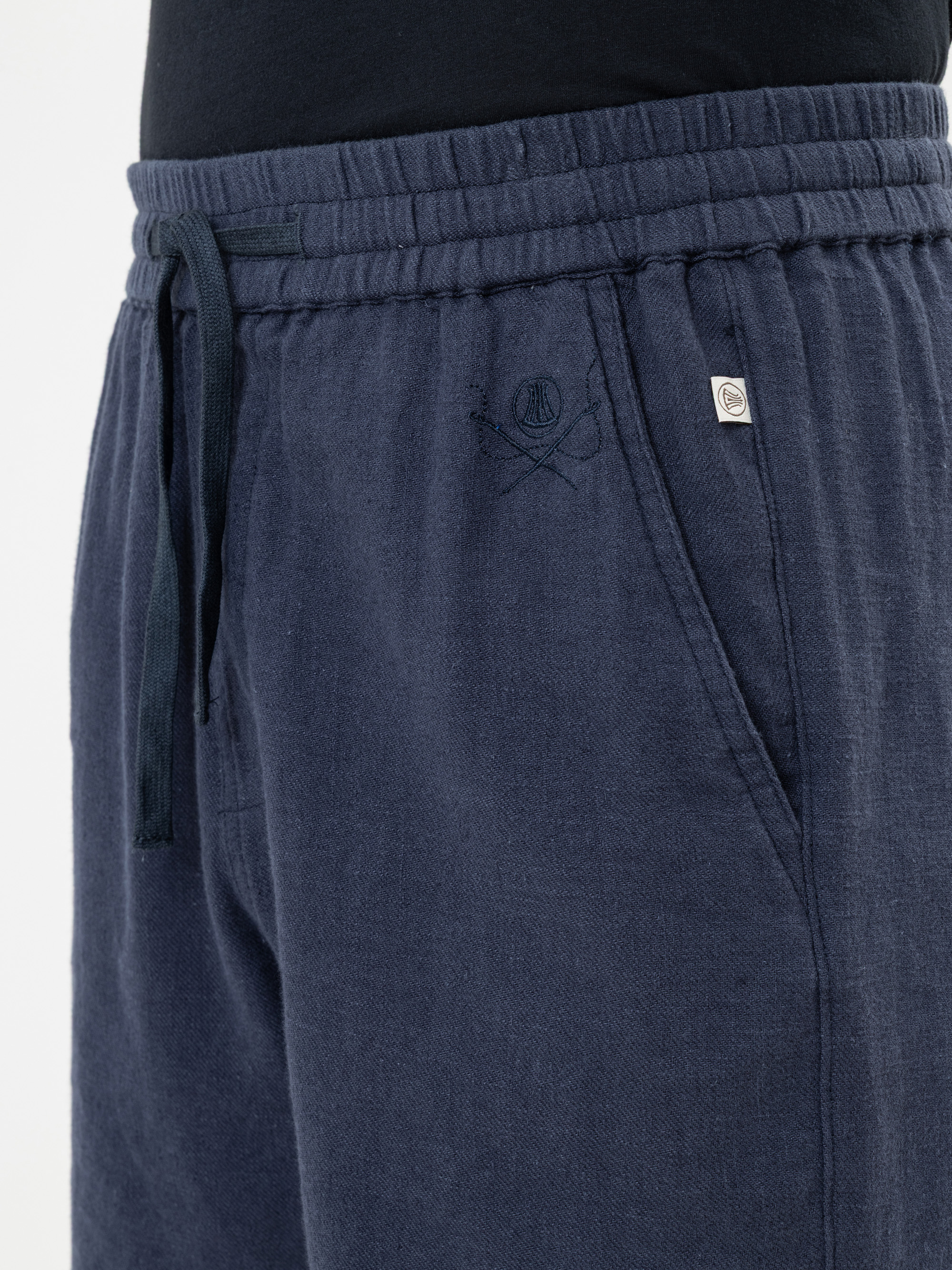 Nahaufnahme einer Person, die eine dunkelblaue Jogginghose mit Kordelzug trägt, die ein gesticktes Logo und einen kleinen Metallknopf in der Nähe der Tasche aufweist. Der Bund ist elastisch, und oben ist ein Teil eines schwarzen Hemdes zu sehen.