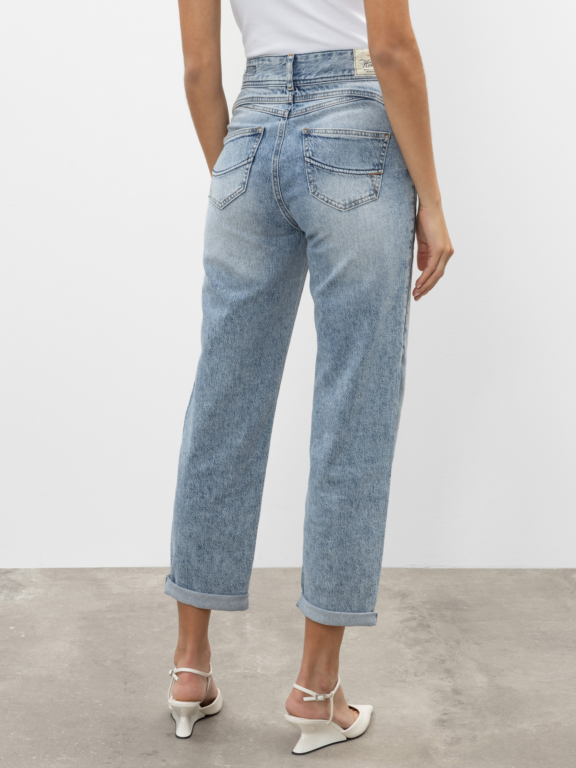 Eine Person steht vor einer schlichten Wand und trägt eine hellblaue Jeans mit hoher Taille und geradem Bein, die Gesäßtaschen hat, sowie weiße Sandalen mit Absatz. Die Jeans sind an den Knöcheln mit Bündchen versehen.