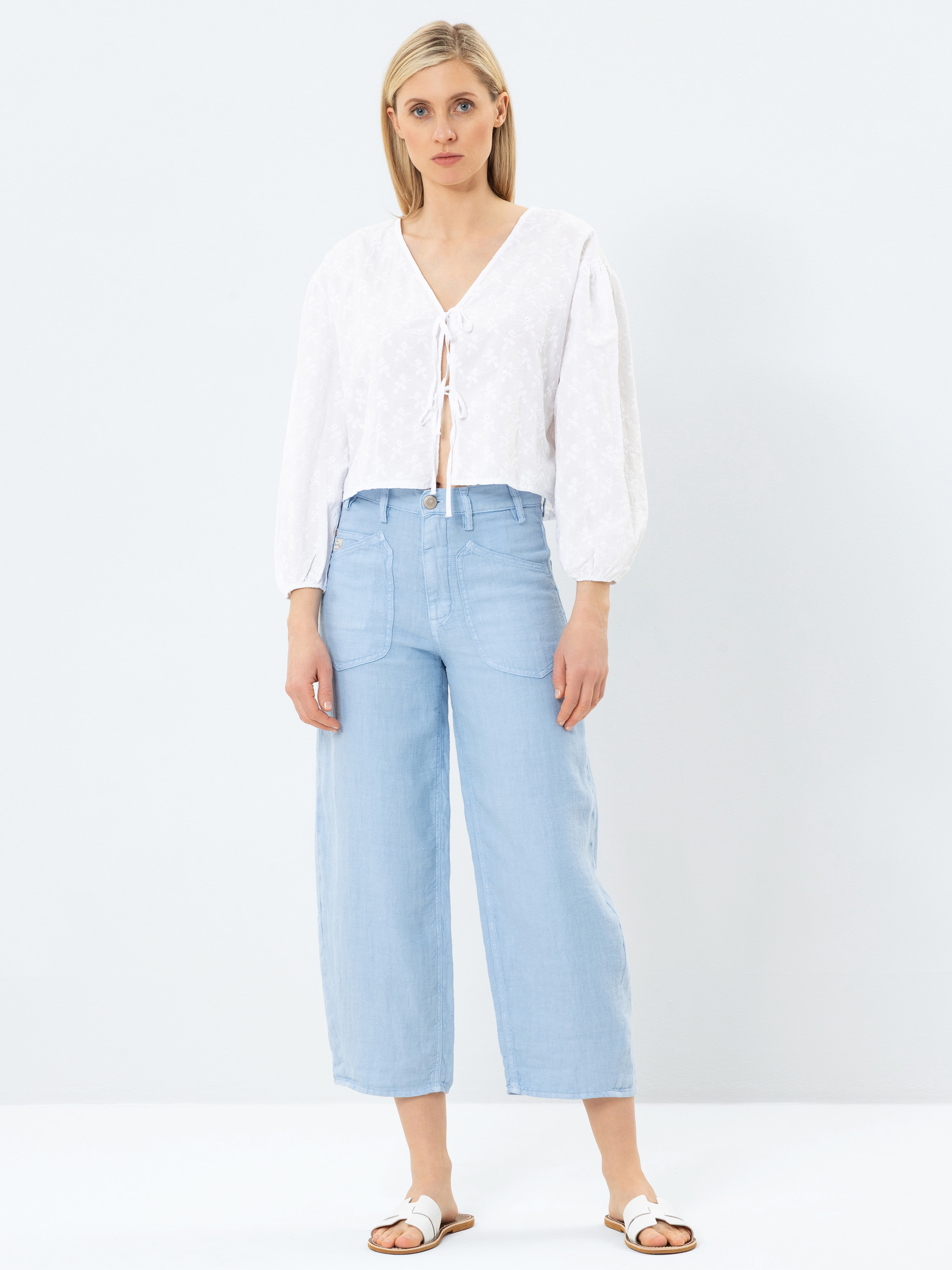 Eine Frau steht vor einem schlichten, hellen Hintergrund. Sie trägt eine weiße, langärmelige Bluse mit Krawatte, hellblaue Jeans mit hoher Taille und weiten Beinen sowie weiße Sandalen. Sie hat glattes blondes Haar und einen neutralen Gesichtsausdruck