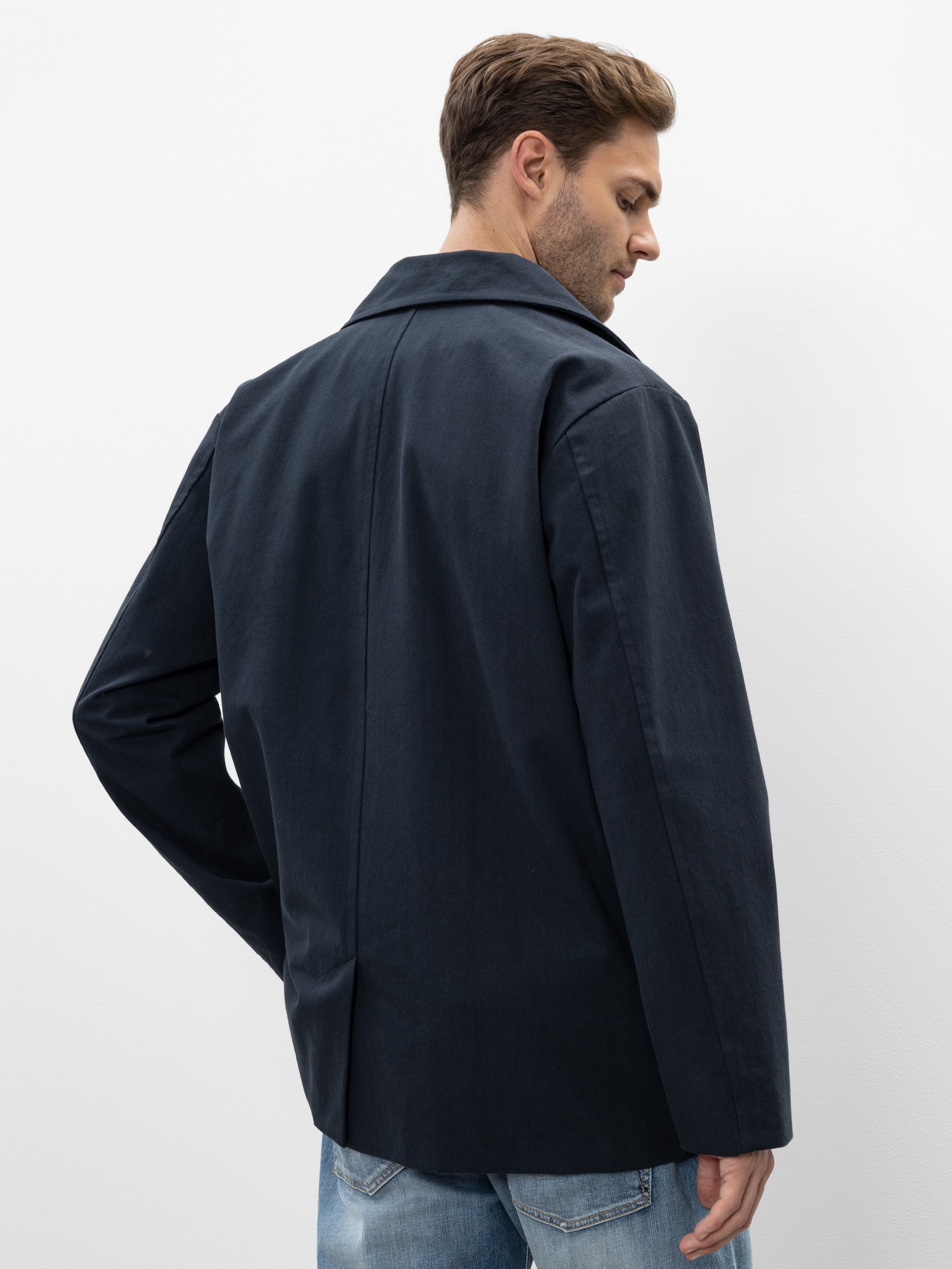 Ein Mann, der einen dunkeln marineblauen Blazer und hellblaue Jeans trägt, steht mit dem Rücken zur Kamera, den Kopf leicht zur Seite gedreht, vor einem schlichten weißen Hintergrund.