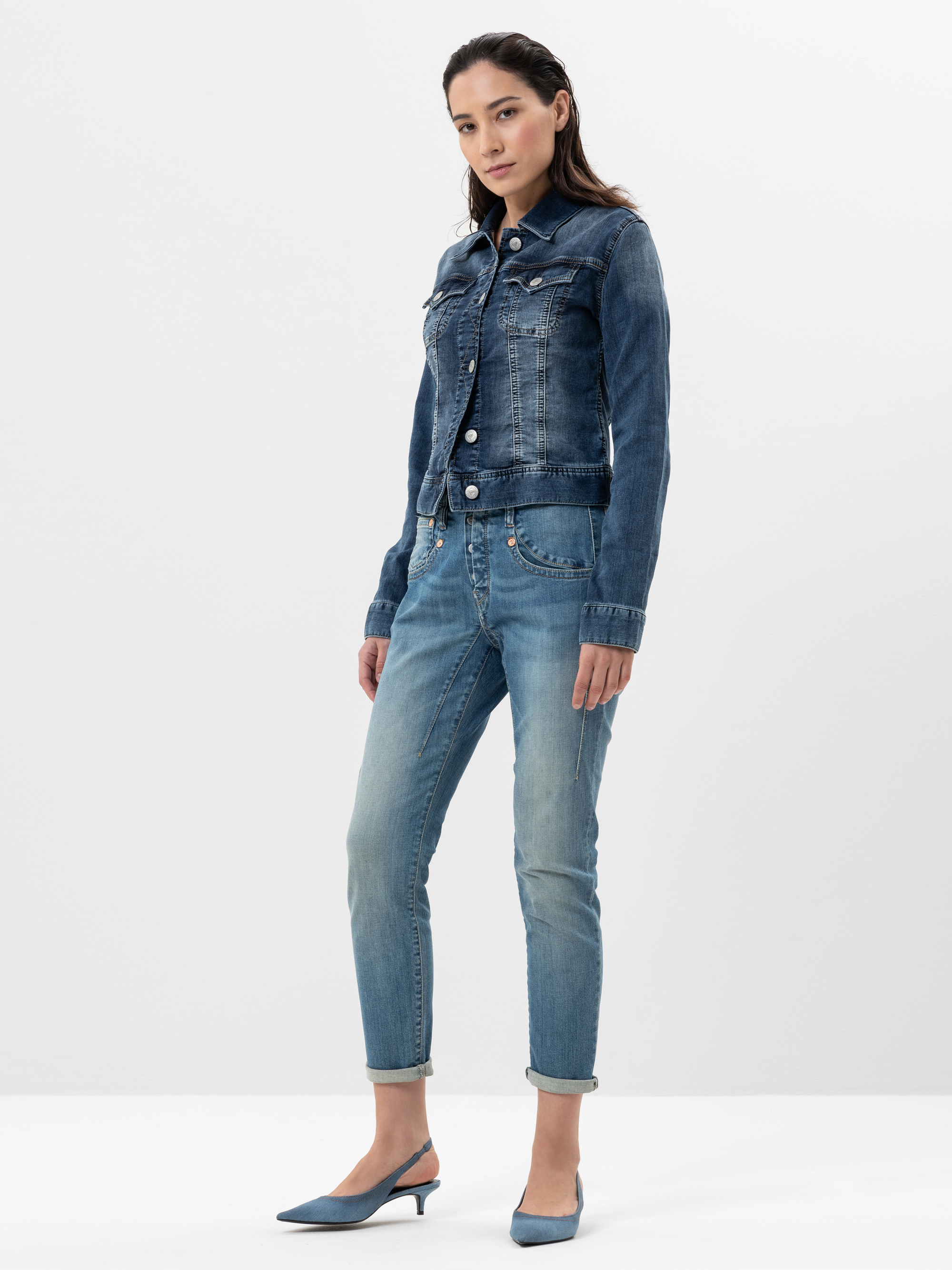 Eine Frau steht vor einem schlichten weißen Hintergrund und trägt eine blaue Jeansjacke, dazu passende blaue Jeans und hellblaue Schuhe mit Absätzen. Sie hat langes dunkles Haar und einen neutralen Gesichtsausdruck, die Hände sind an den Seiten versch