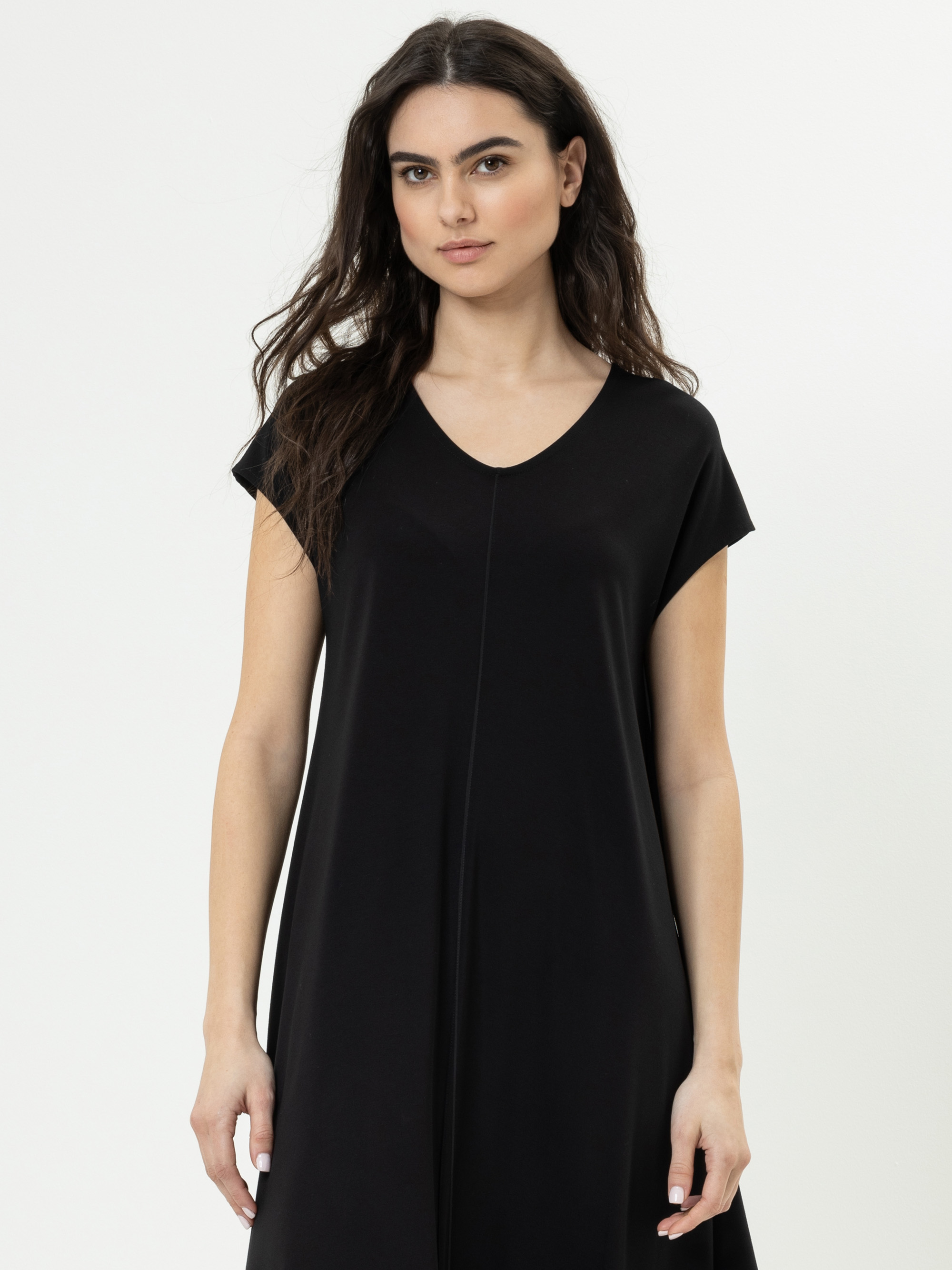 Eine Frau mit langen, gewellten braunen Haaren trägt ein kurzärmeliges schwarzes Kleid mit V-Ausschnitt, steht vor einem schlichten weißen Hintergrund und schaut mit neutralem Blick in die Kamera.