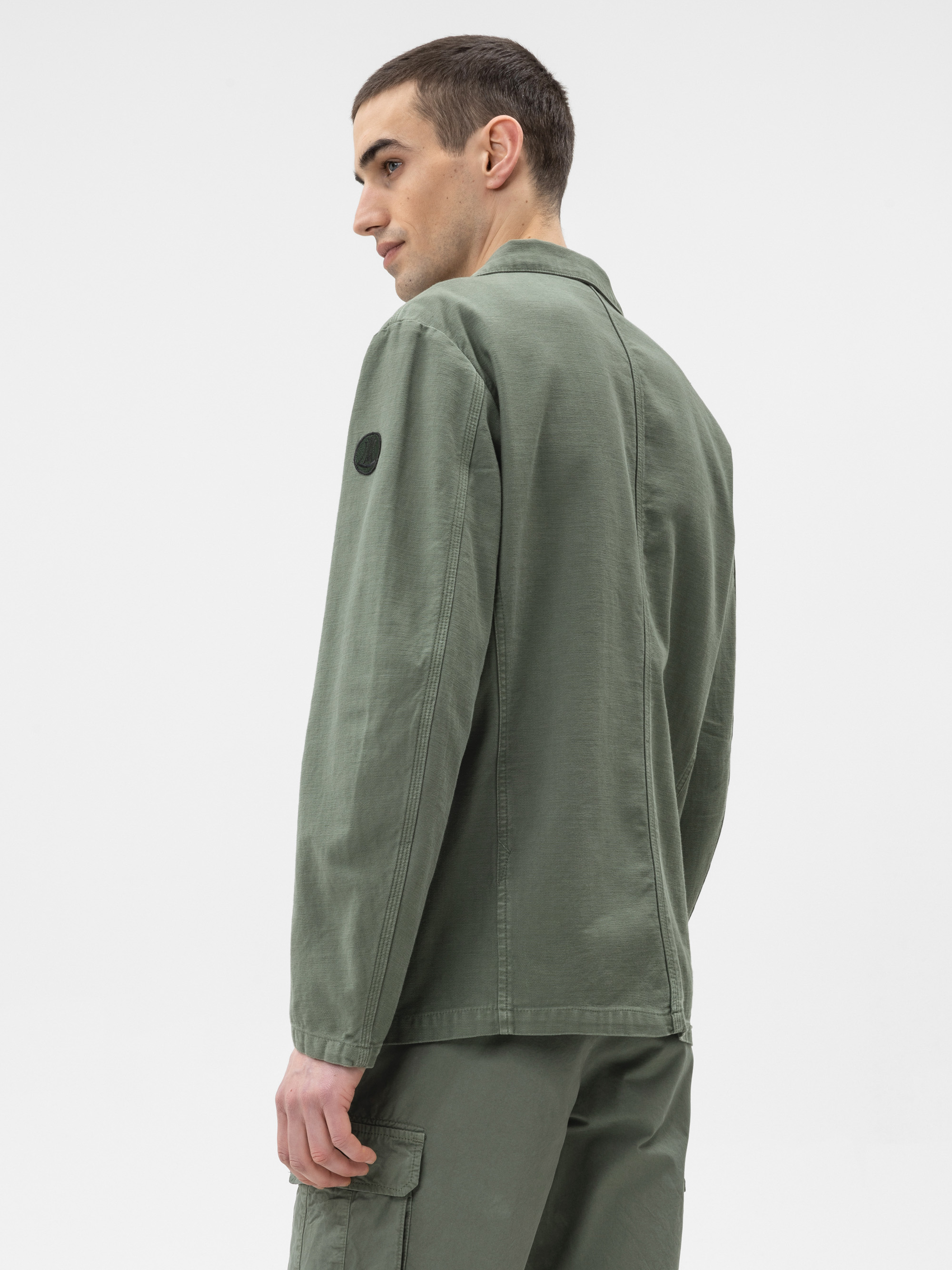 Eine Person in einer olivgrünen Arbeitsjacke und einer dazu passenden Hose steht leicht zur Seite gedreht vor einem schlichten weißen Hintergrund und schaut über ihre Schulter.