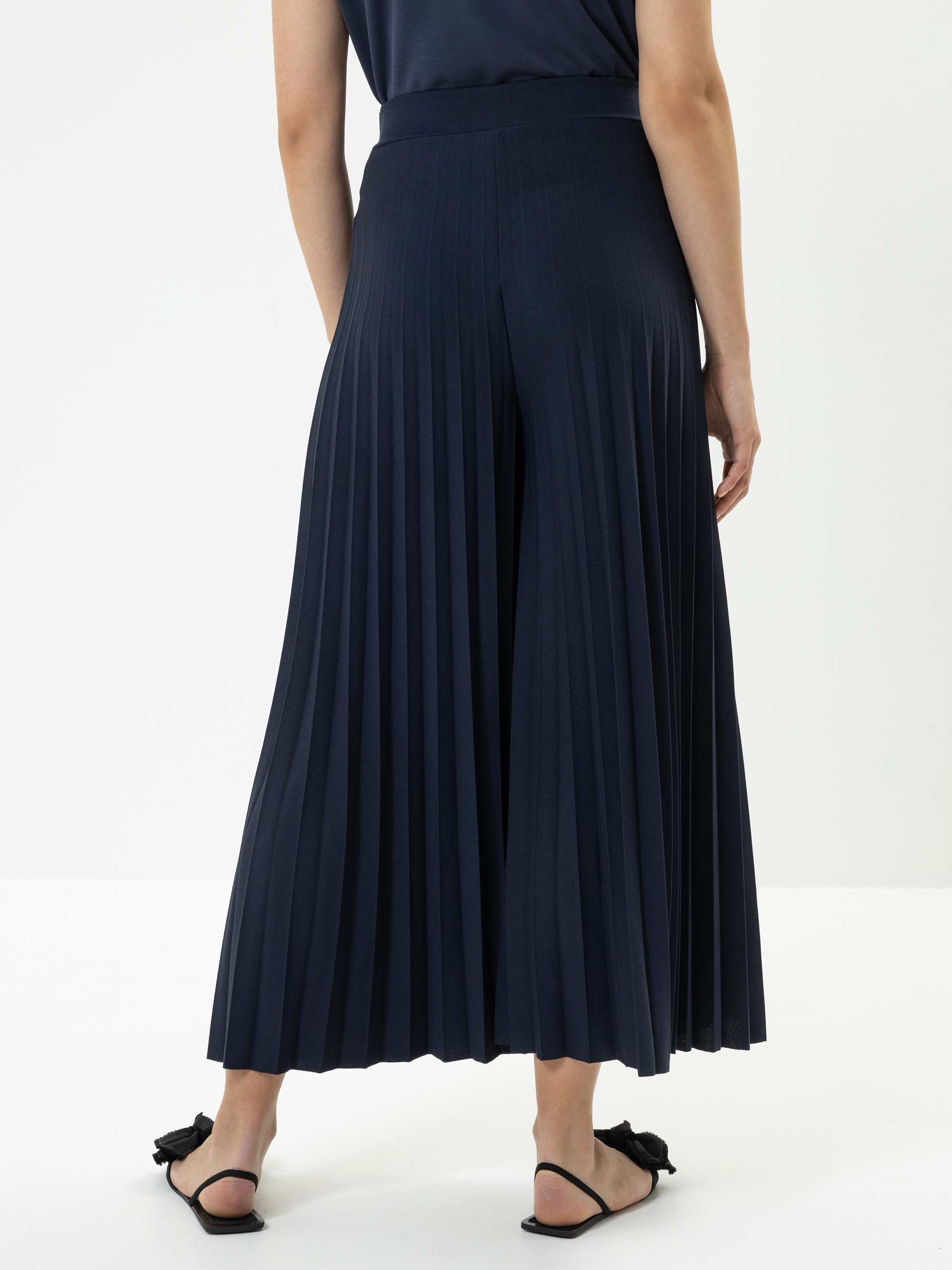 Eine Person steht mit dem Gesicht zur Wand, trägt eine marineblaue, plissierte Hose mit weitem Bein und schwarze Sandalen mit Schleifen vor einem schlichten weißen Hintergrund.