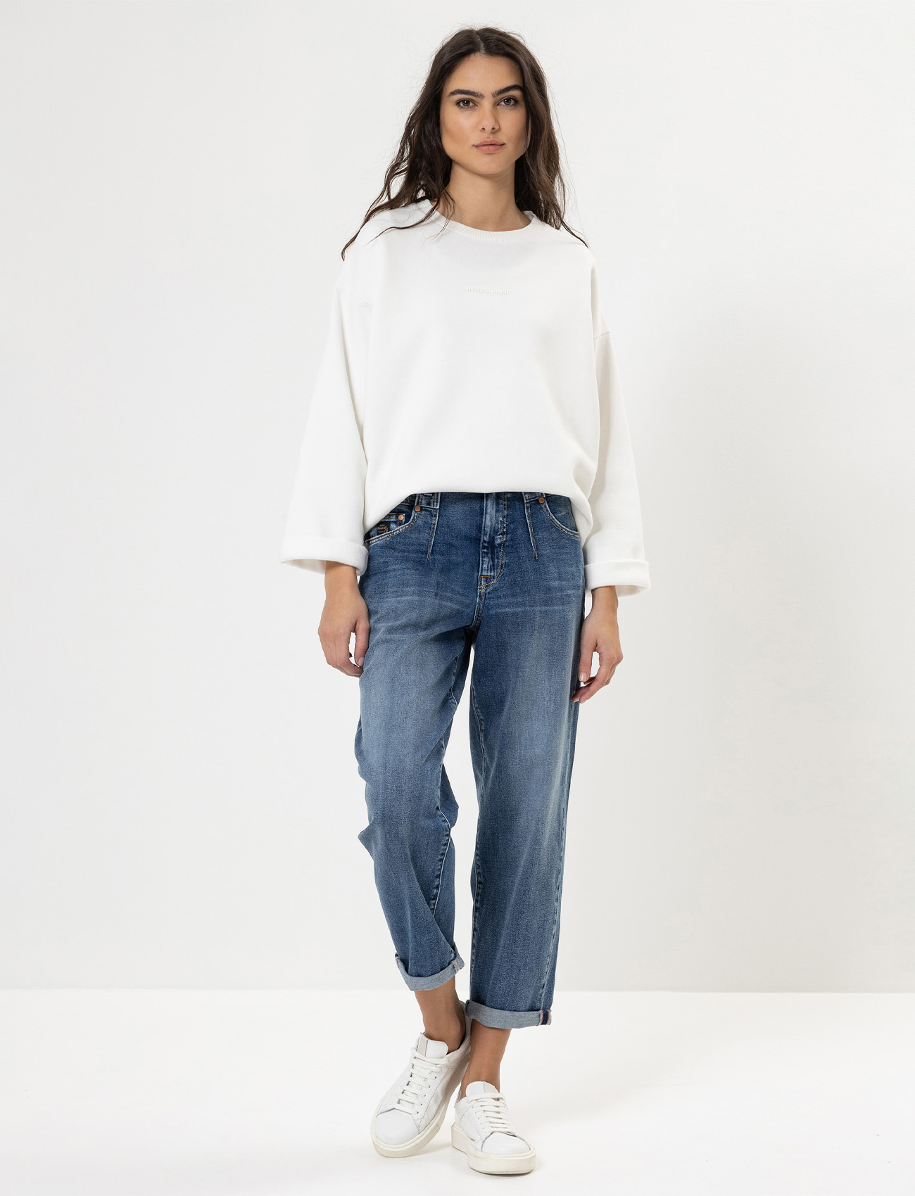 Eine Frau mit langen dunklen Haaren trägt ein lockeres weißes Sweatshirt, blaue Jeans mit Bündchen und weiße Turnschuhe und steht vor einem schlichten weißen Hintergrund.