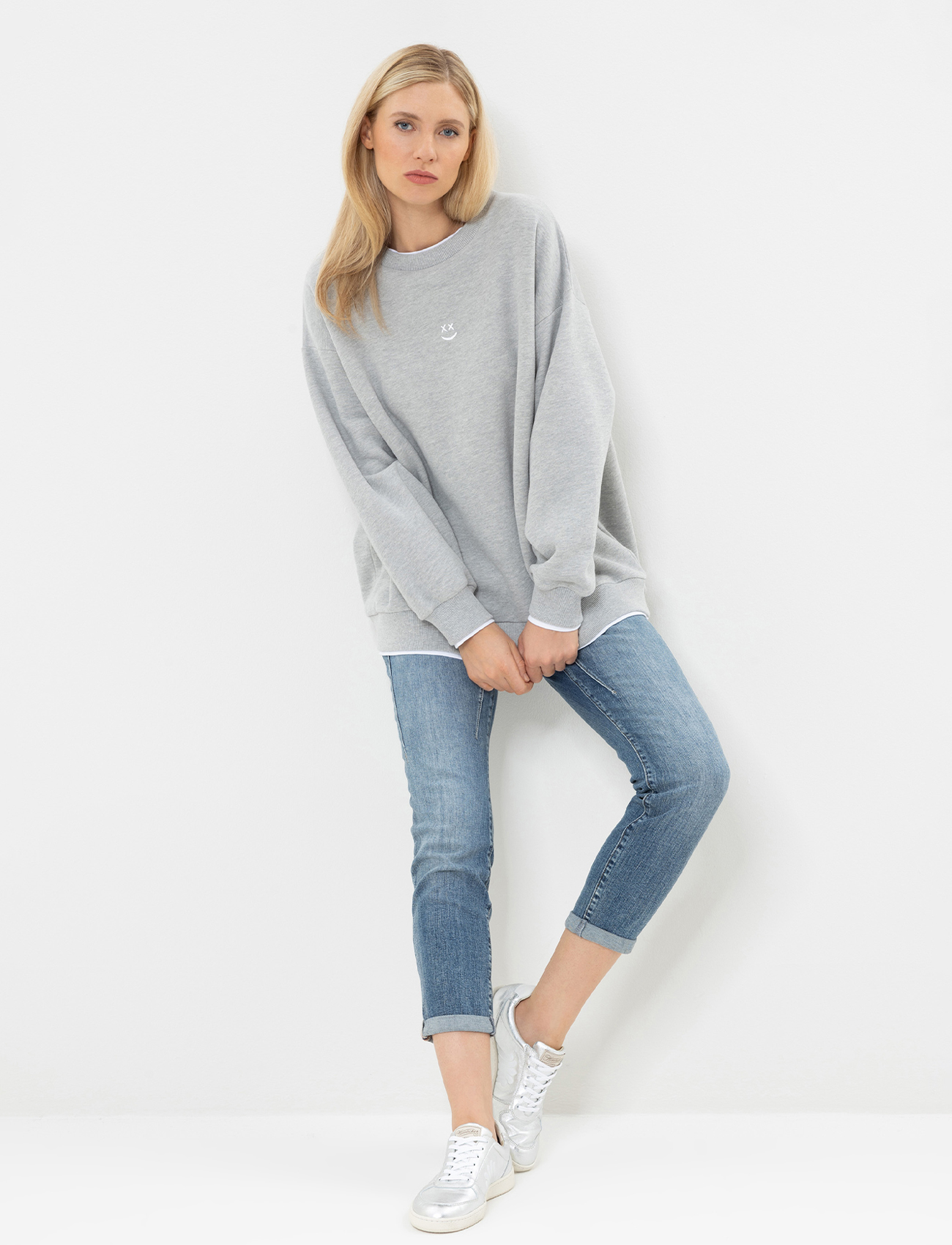 Eine Frau mit langen dunklen Haaren trägt ein lockeres weißes Sweatshirt, blaue Jeans mit Bündchen und weiße Turnschuhe und steht vor einem schlichten weißen Hintergrund.