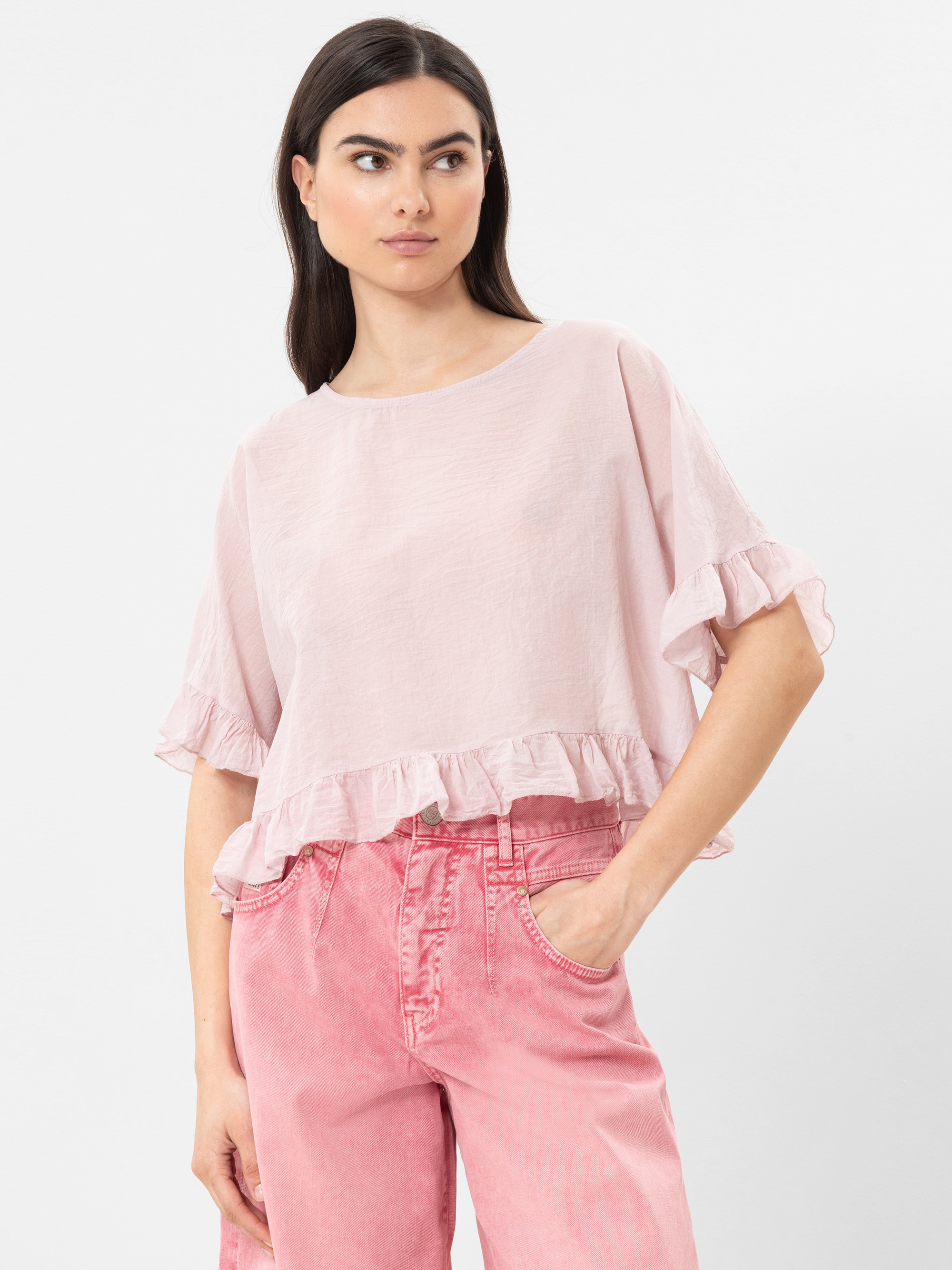 Frau mit langen dunklen Haaren, die eine hellrosa, kurzärmelige Rüschenbluse und hochgekrempelte rosa Jeans trägt, steht mit einer Hand in der Tasche und blickt zur Seite vor einem schlichten Hintergrund.