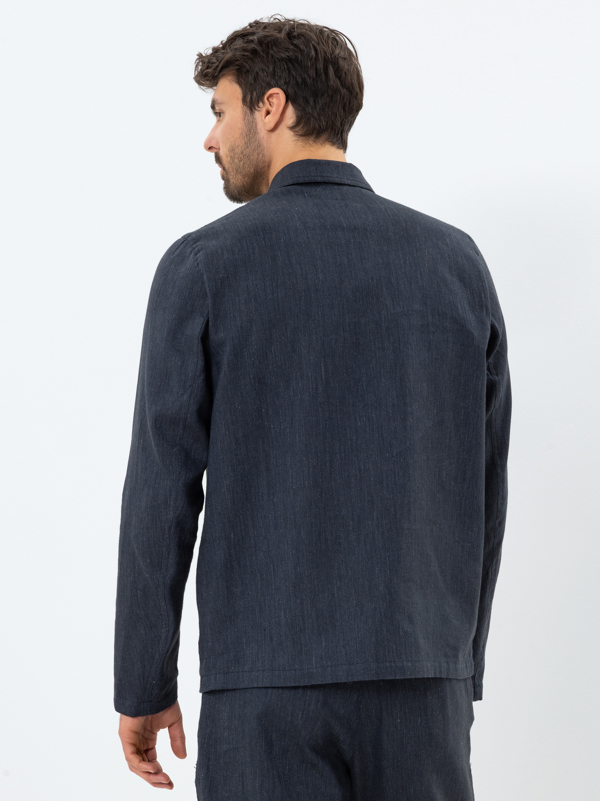Ein Mann mit kurzen braunen Haaren, der ein dunkelblaues langärmeliges Hemd und eine dazu passende Hose trägt, steht mit dem Gesicht zur Kamera vor einem schlichten weißen Hintergrund.