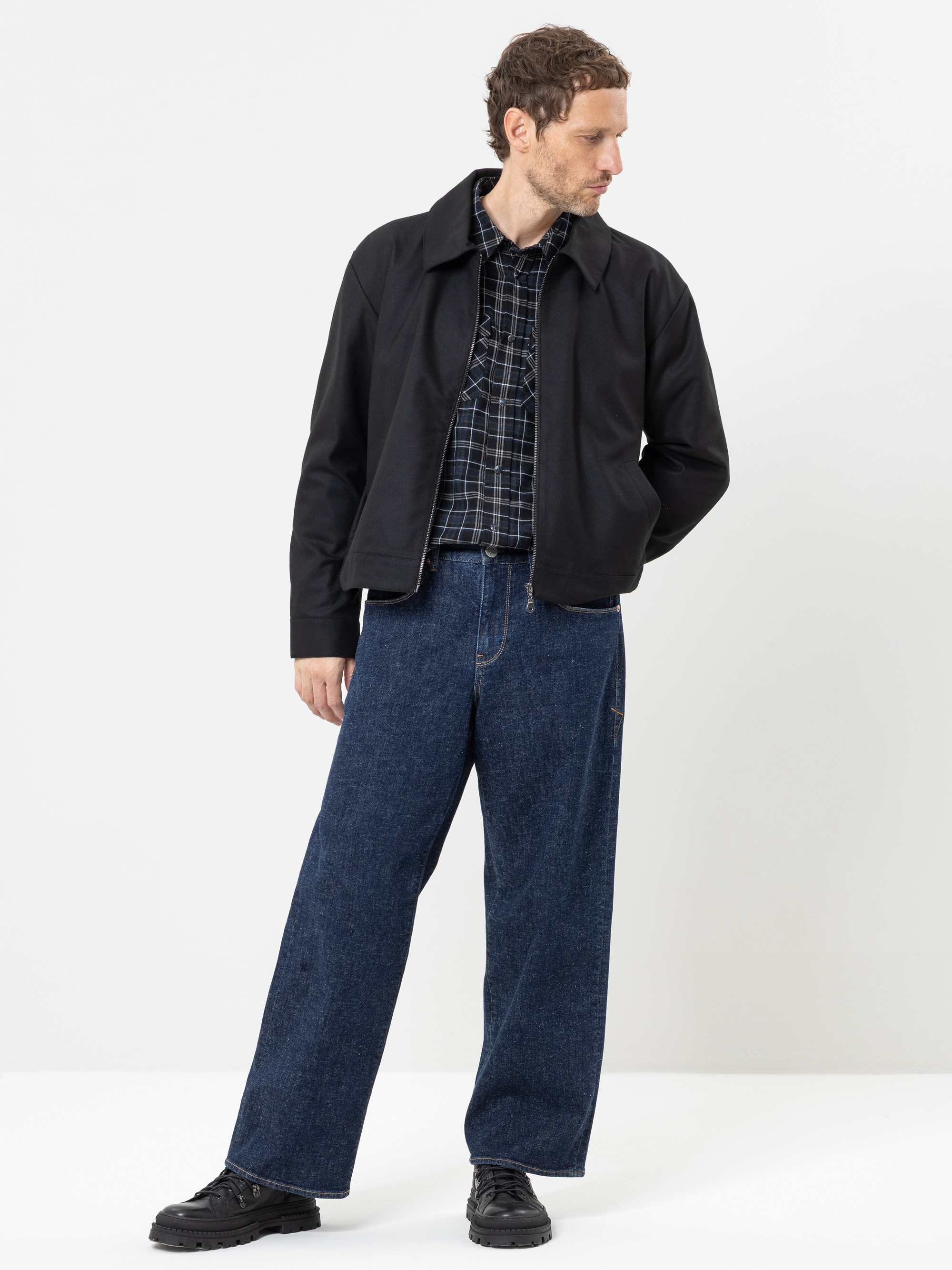 Ein Mann, der eine schwarze Jacke, ein kariertes Hemd, eine blaue Jeans mit weitem Bein und schwarze Schuhe trägt, steht vor einem schlichten weißen Hintergrund und blickt nach links.