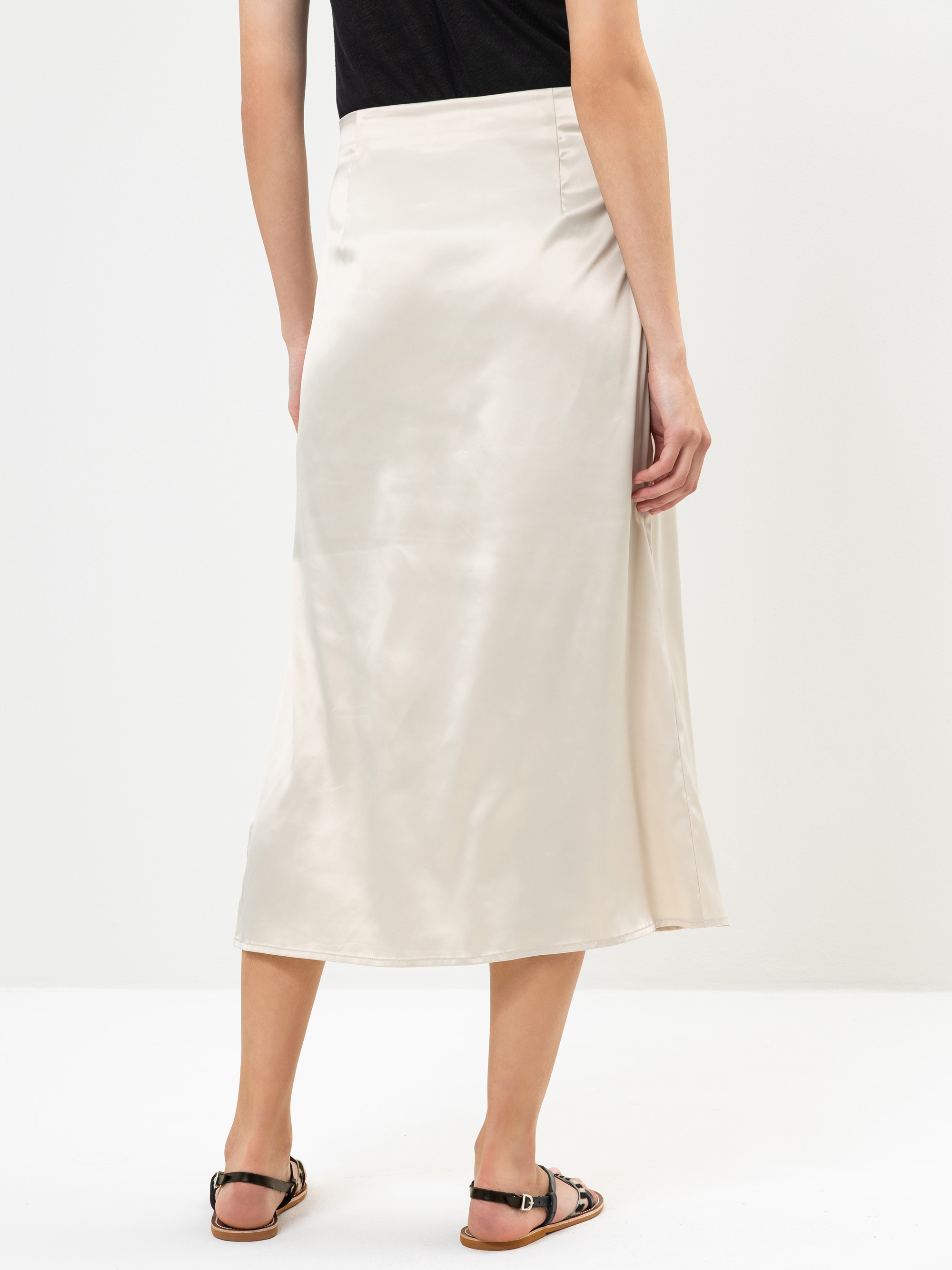 Eine Person, die ein schwarzes Oberteil, einen hellbeigen Midirock aus Satin und schwarze Sandalen trägt, steht mit dem Rücken zur Kamera vor einem schlichten weißen Hintergrund.