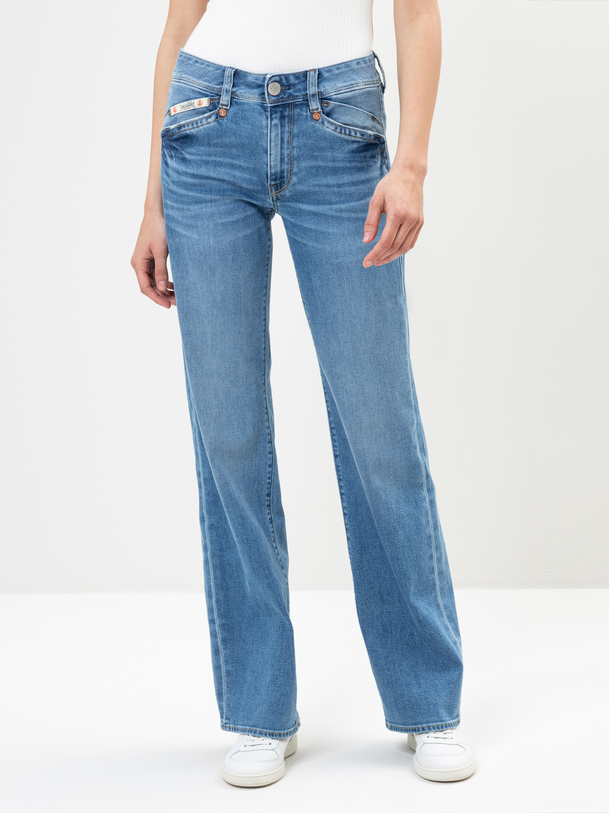 Person in hellblauen, weit geschnittenen Jeans und einem weißen, gerippten Oberteil, die vor einem schlichten, hellen Hintergrund steht. Die Jeans hat Vordertaschen und die Person trägt außerdem weiße Turnschuhe.