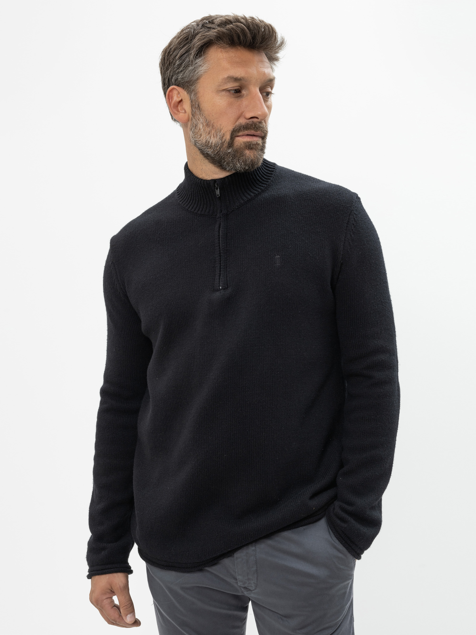 Ein Mann mit kurzen, dunklen Haaren und einem Bart trägt einen schwarzen Pullover mit Viertelreißverschluss und eine graue Hose. Er steht vor einem schlichten weißen Hintergrund und schaut nach links.