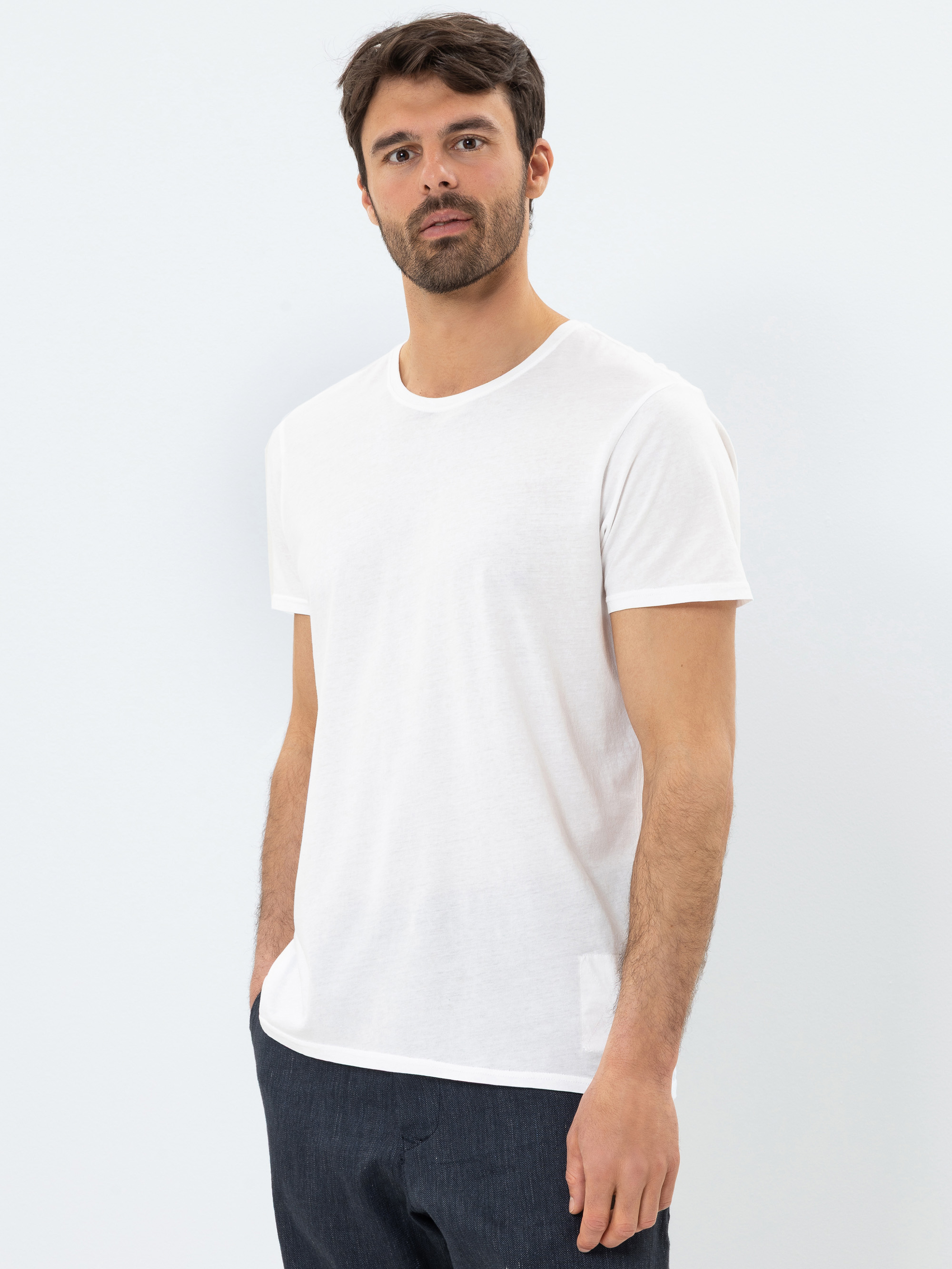 Ein Mann mit kurzen dunklen Haaren und einem Bart trägt ein schlichtes weißes T-Shirt und eine dunkle Hose und steht mit neutralem Gesichtsausdruck vor einem hellen Hintergrund.