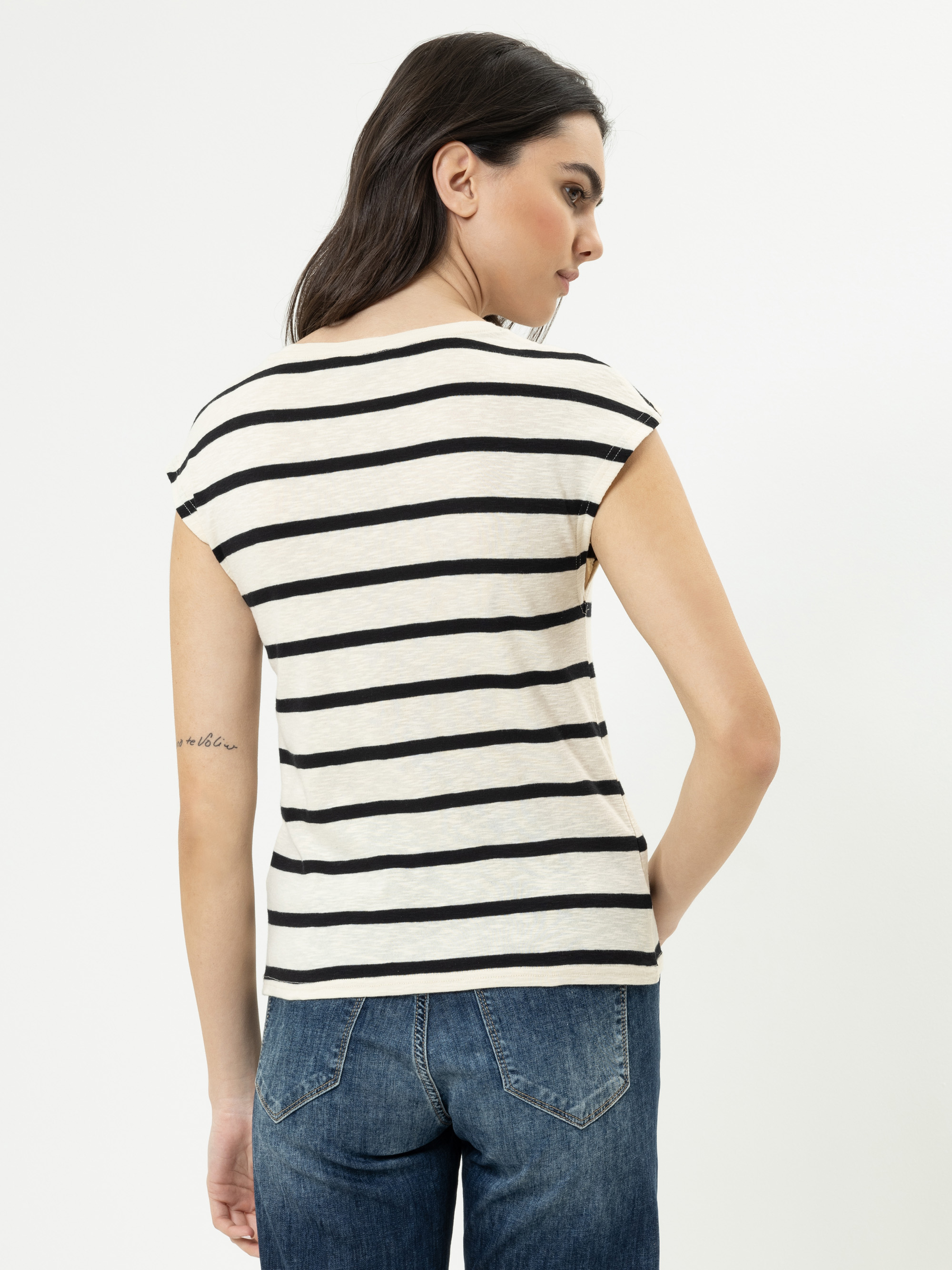 Eine Frau mit langen dunklen Haaren, die ein kurzärmeliges, schwarz-weiß gestreiftes Oberteil und blaue Jeans trägt, steht mit dem Rücken zur Kamera vor einem schlichten weißen Hintergrund.