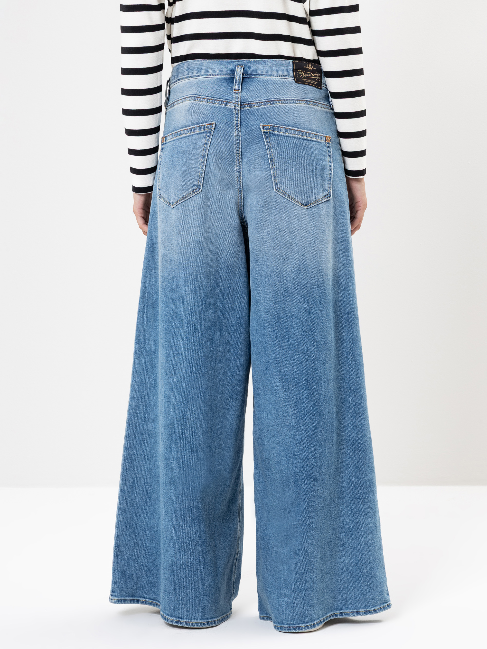 Eine Person, die ein schwarz-weiß gestreiftes langärmeliges Hemd und eine weit geschnittene blaue Jeans trägt, ist von hinten zu sehen und steht vor einem schlichten weißen Hintergrund.