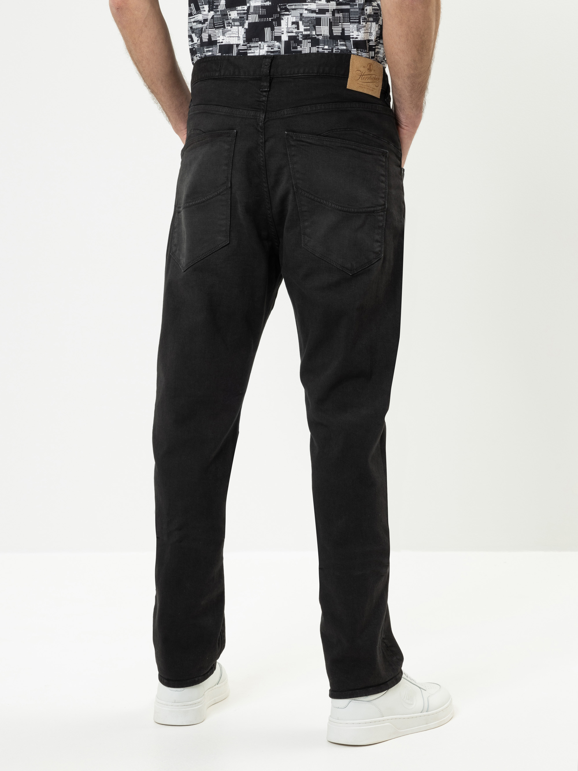 Eine Person, die eine schwarze Jeans und weiße Turnschuhe trägt, steht mit dem Gesicht nach hinten. Die Jeans hat zwei Gesäßtaschen und einen hellbraunen Aufnäher am Bund. Die Person trägt außerdem ein gemustertes schwarz-weißes Hemd.