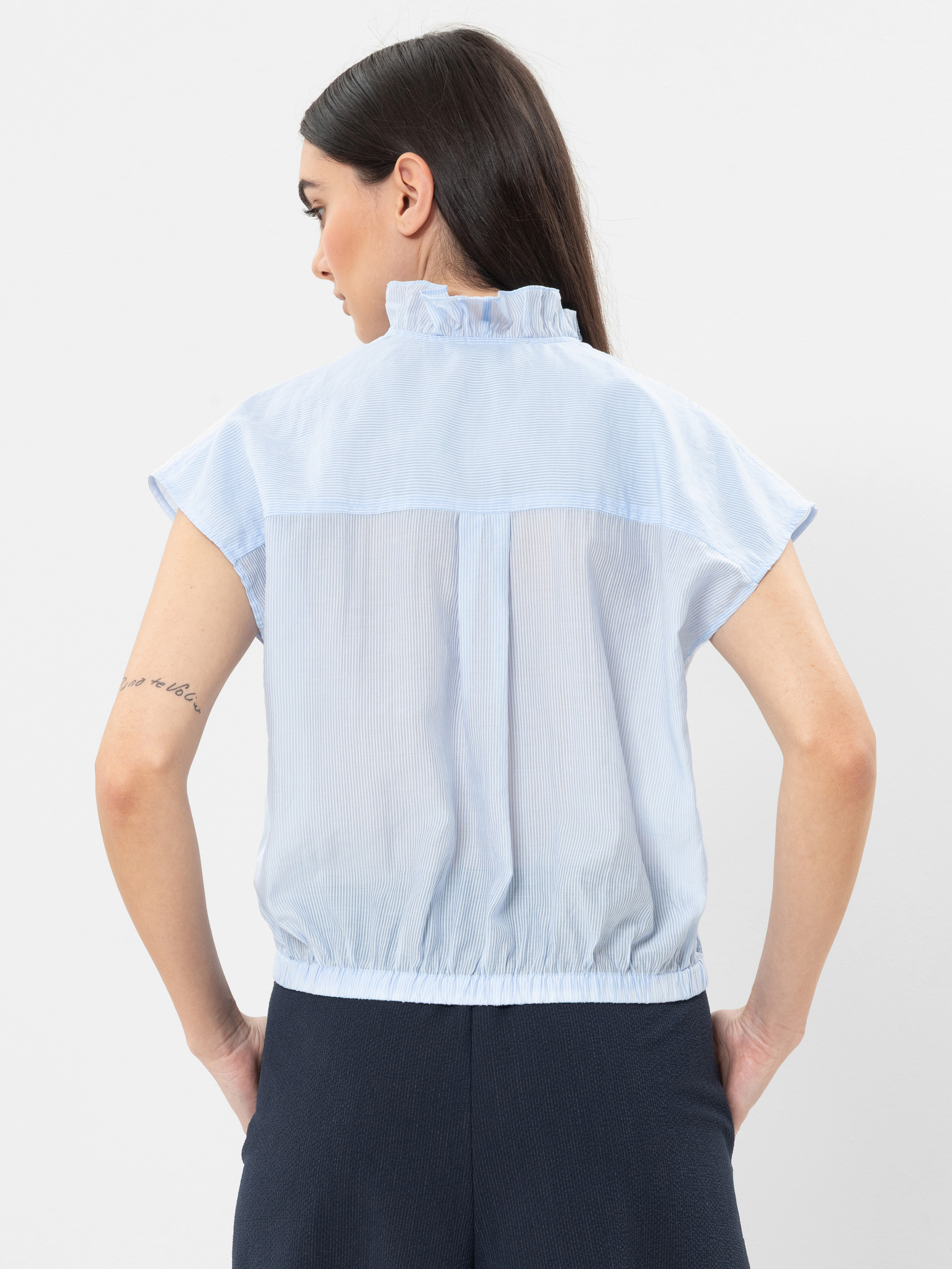 Eine Frau mit langen dunklen Haaren, von hinten gesehen, trägt eine hellblaue kurzärmelige Bluse mit Rüschenkragen und eine dunkle Hose. Ihre Arme sind an den Ellbogen angewinkelt und sie hat eine Tätowierung auf dem linken Unterarm.