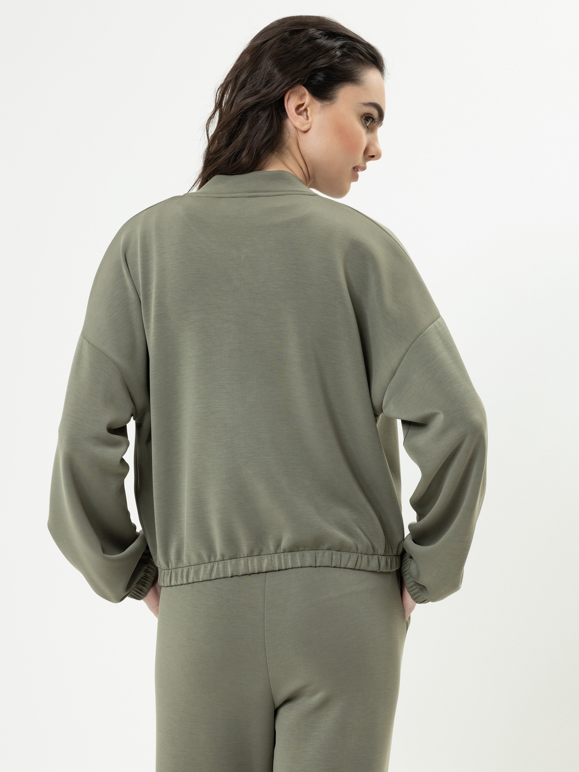 Eine Frau mit langen dunklen Haaren steht mit dem Rücken zur Kamera in einem locker sitzenden, olivgrünen Trainingsanzug mit den Händen in den Taschen vor einem schlichten weißen Hintergrund.
