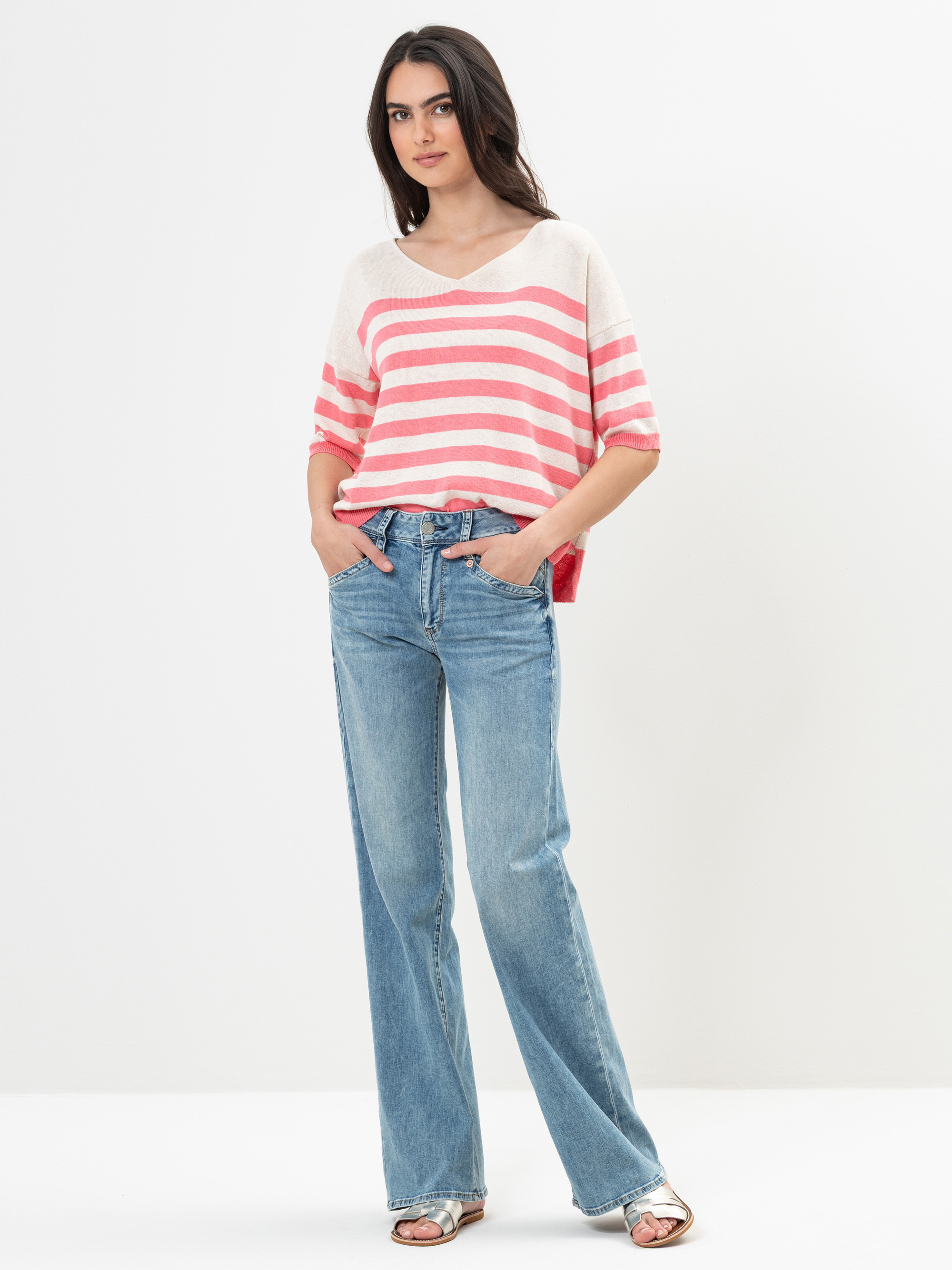 Eine Frau steht vor einem weißen Hintergrund, trägt ein cremefarbenes und rosa gestreiftes kurzärmeliges Oberteil, hellblaue Jeans mit weitem Bein und Sandalen, die Hände in den Taschen.